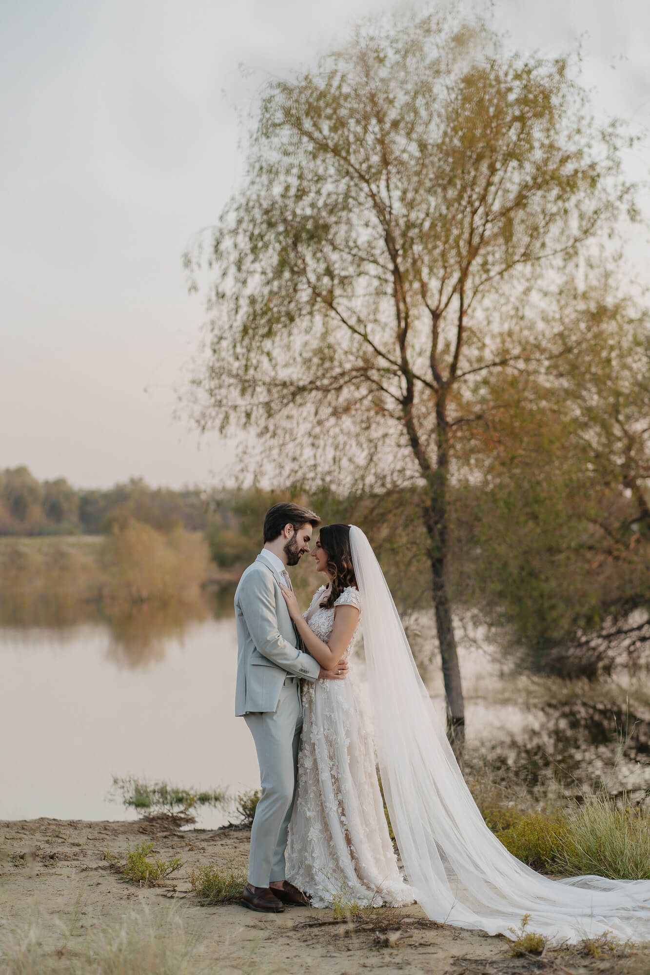 A couple in wedding attire shares romantic moments by a lake with autumn trees in the background.