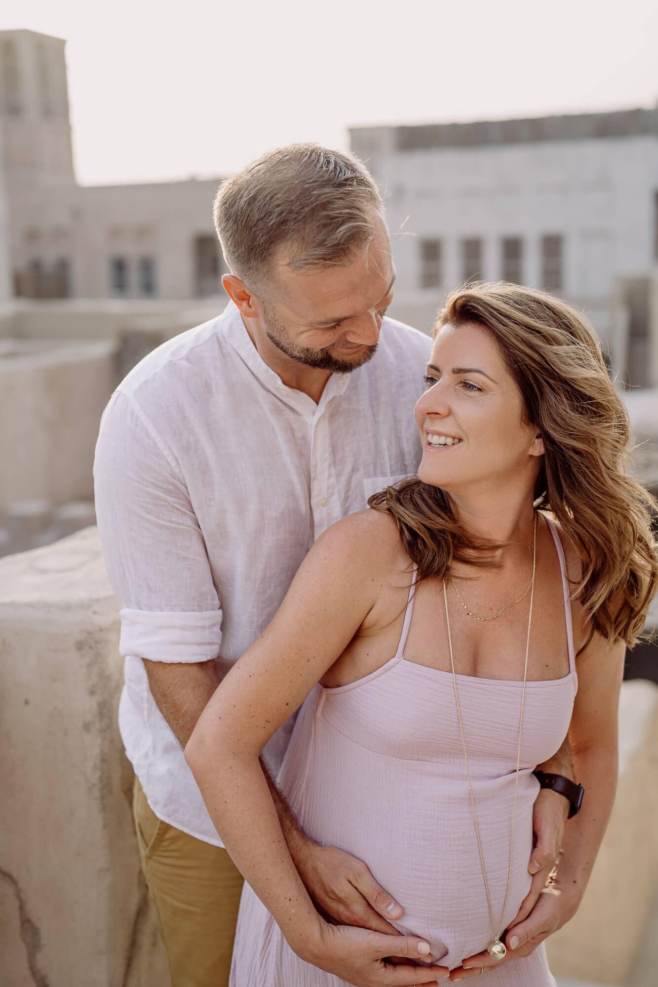 Urban rooftop maternity session featuring couple in soft pink and white against city backdrop.