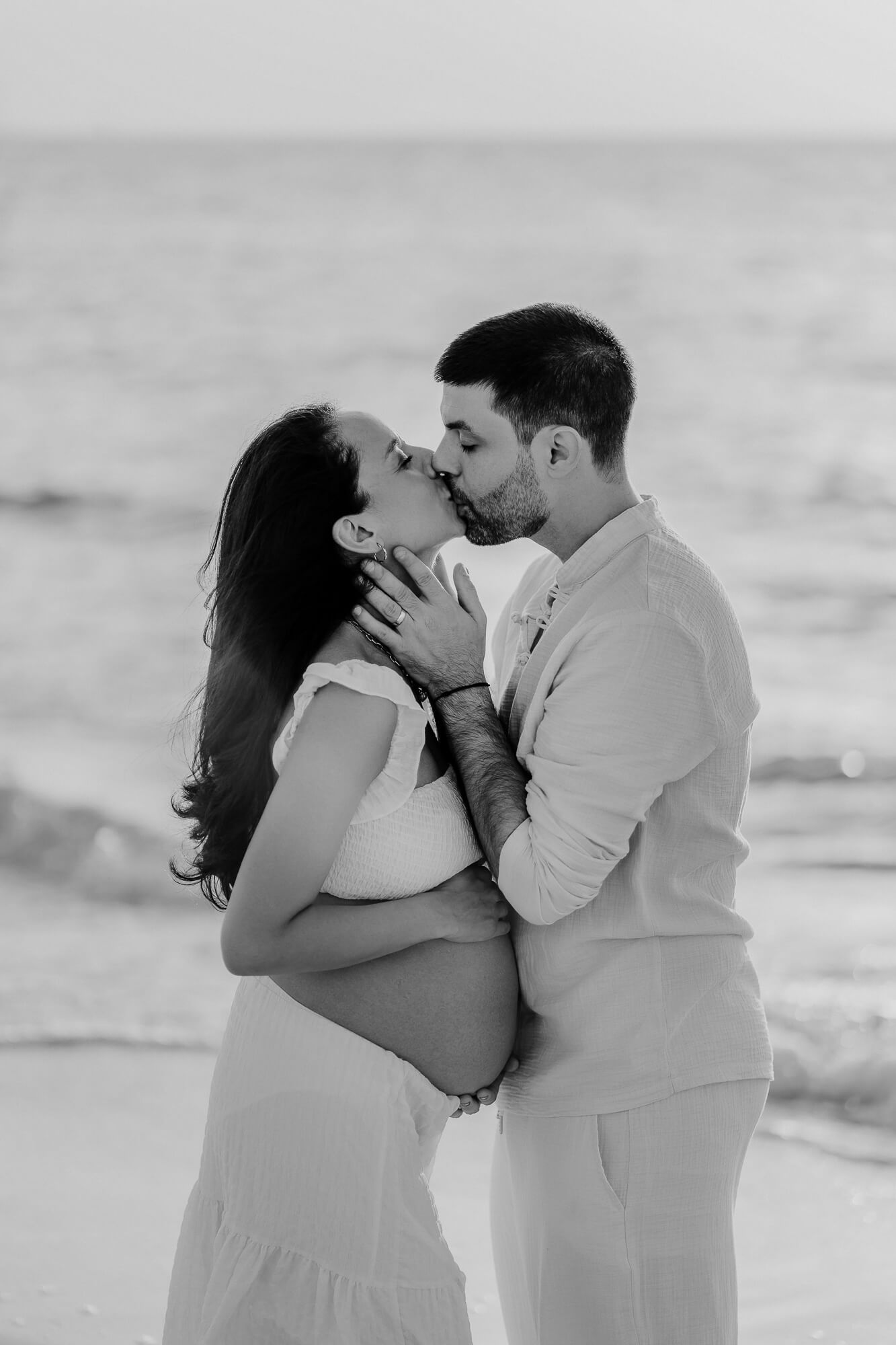 A couple in white outfits sharing romantic moments on the beach during a sunset photoshoot.
