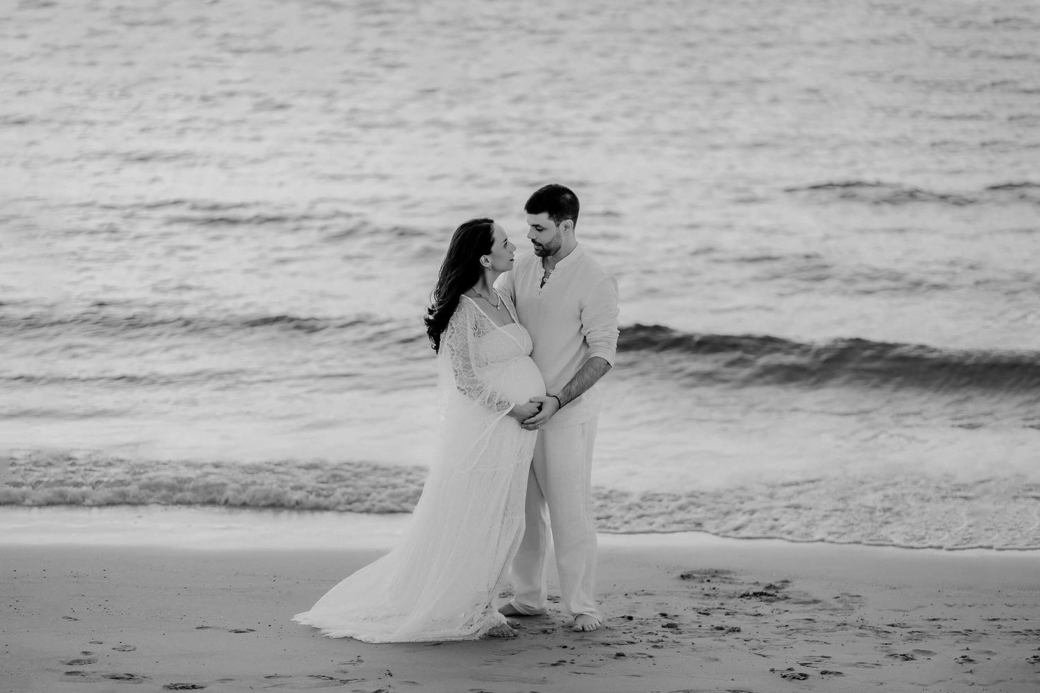 A couple in wedding attire shares an intimate moment on a black and white beach at sunset.