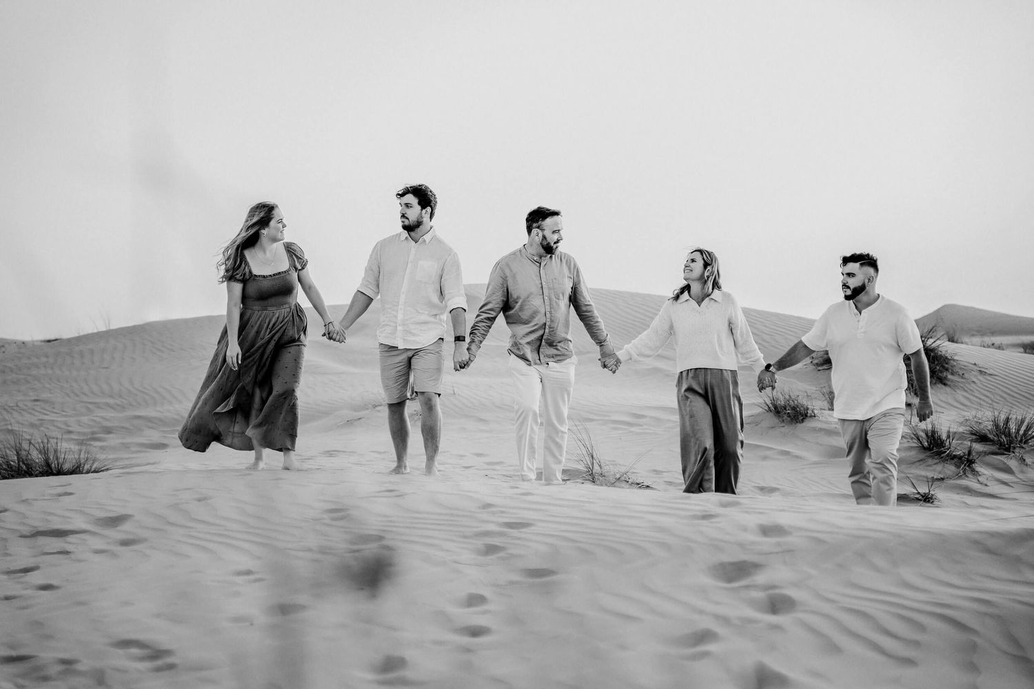 Black and white photo of five people holding hands while walking through desert sand dunes.