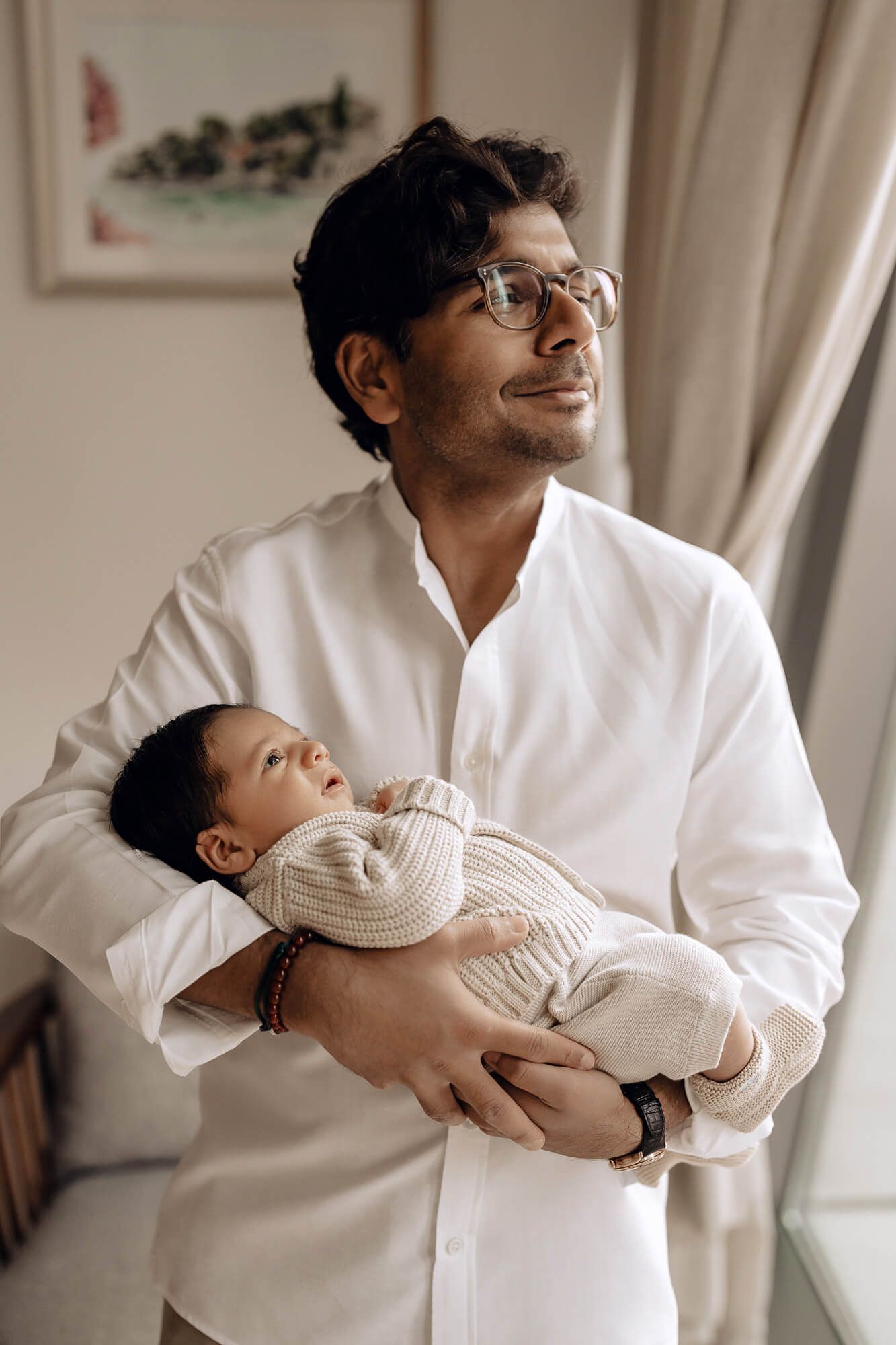 Parent in white shirt gazes lovingly while holding sleeping infant in cream sweater.