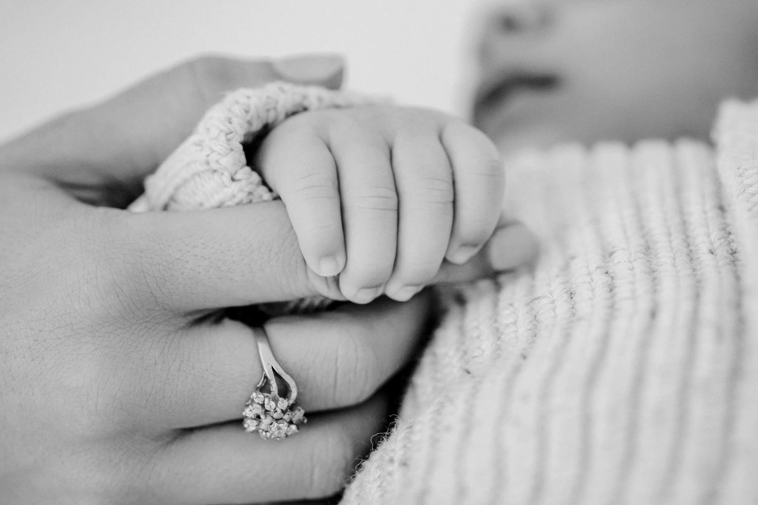 Black and white photo of baby's hand holding parent's finger with ring.