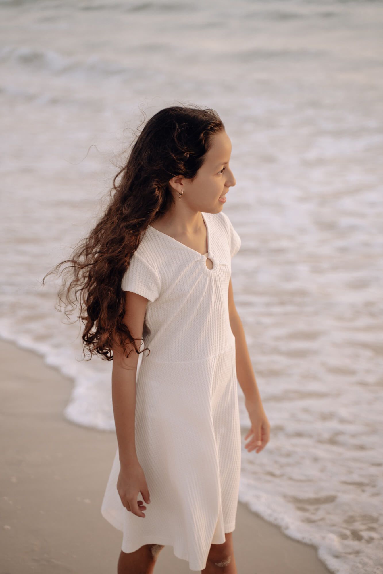 A person with long wavy hair wearing a white dress stands thoughtfully by the ocean.