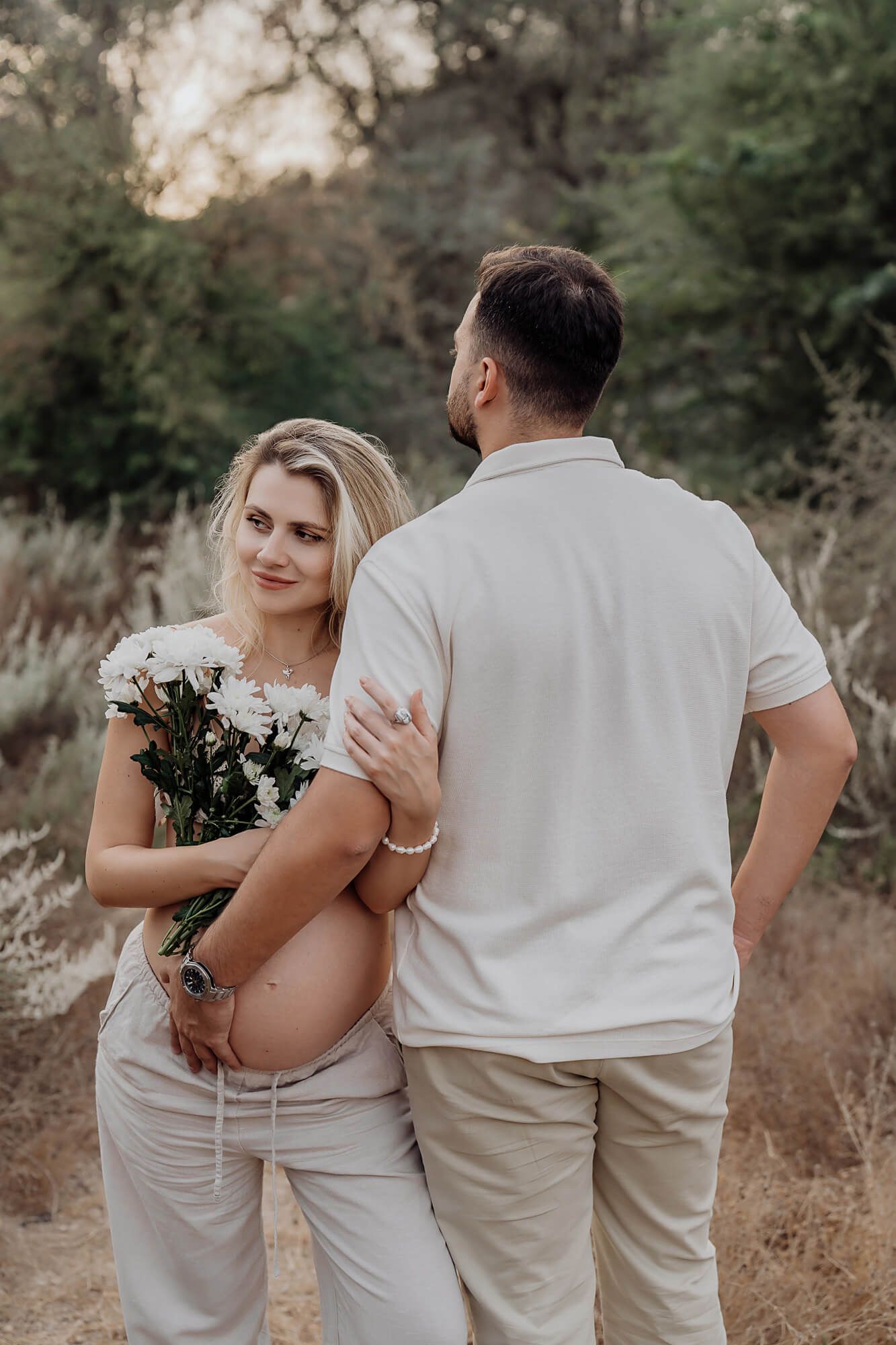 Outdoor maternity photoshoot featuring couple in light-colored clothing holding white flowers on a nature trail.