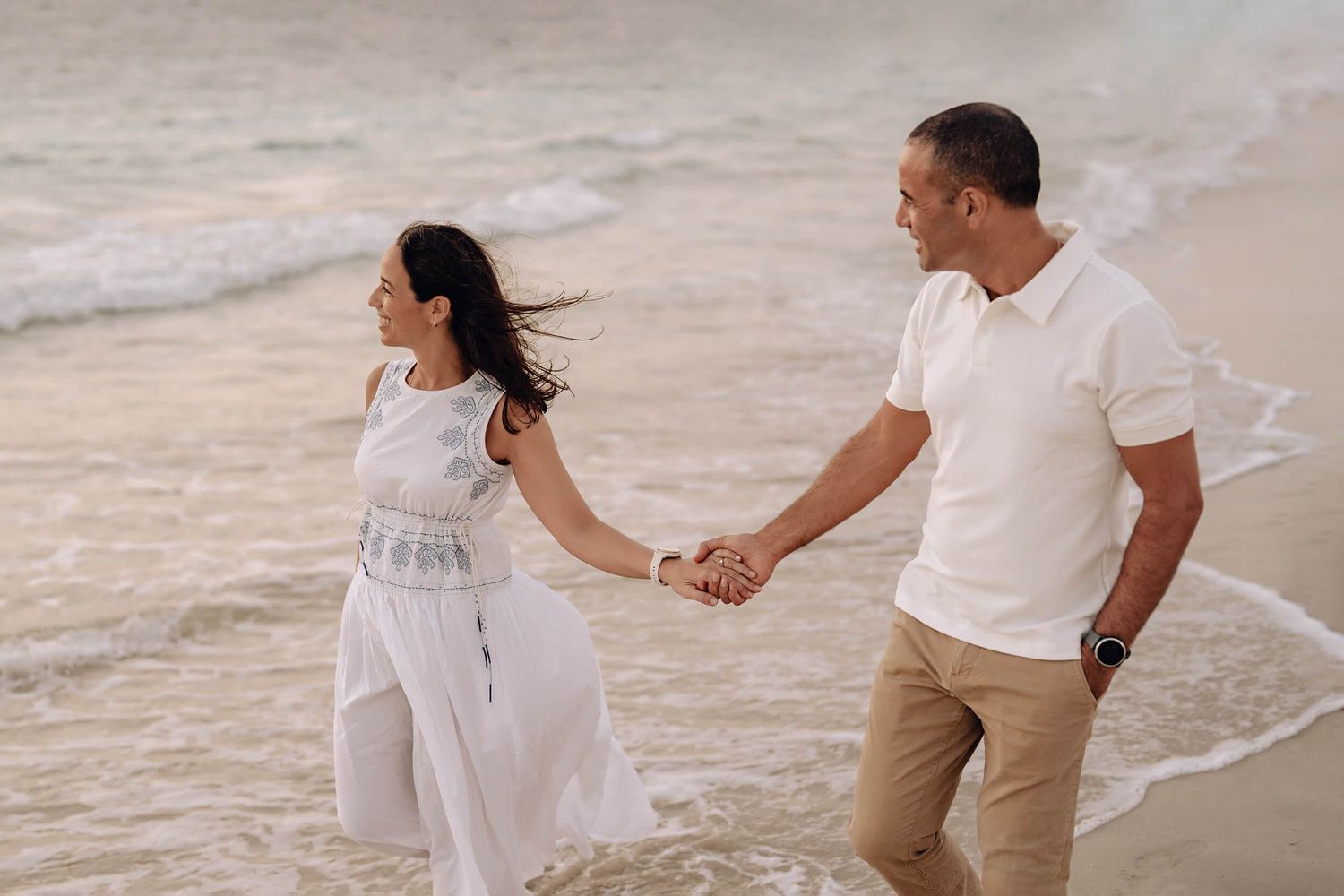 Couple strolling along beach holding hands with wind blowing through their hair.