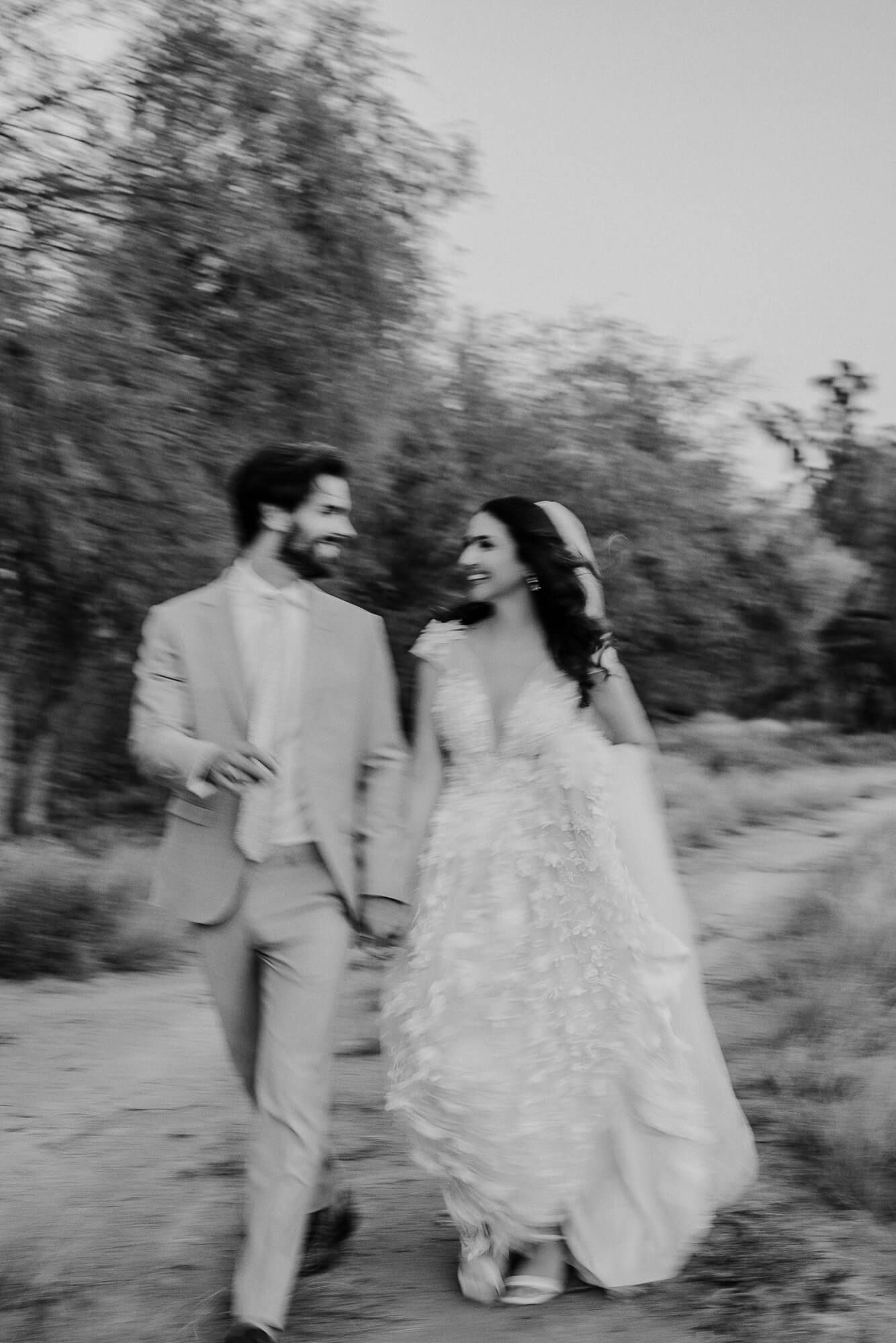 A black and white photo of a bride and groom walking together outdoors, sharing a joyful moment in nature.