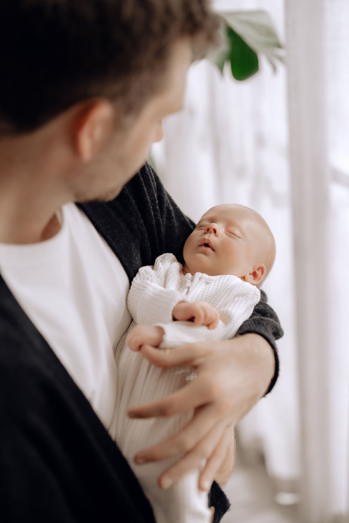 A tender moment between parent and newborn baby dressed in white against bright natural window light.