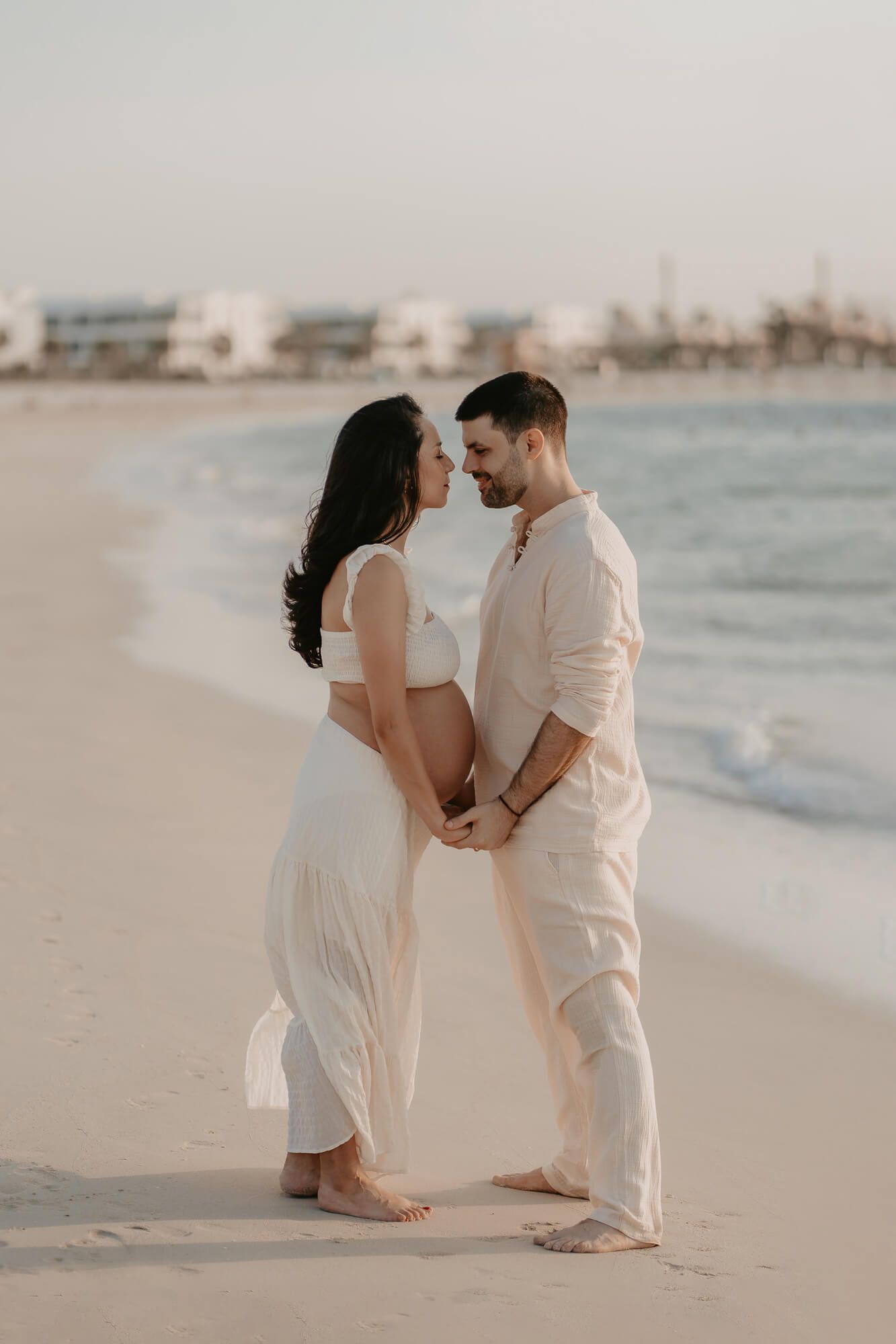 A couple in white attire stands together romantically on a serene beach with soft waves in the background.