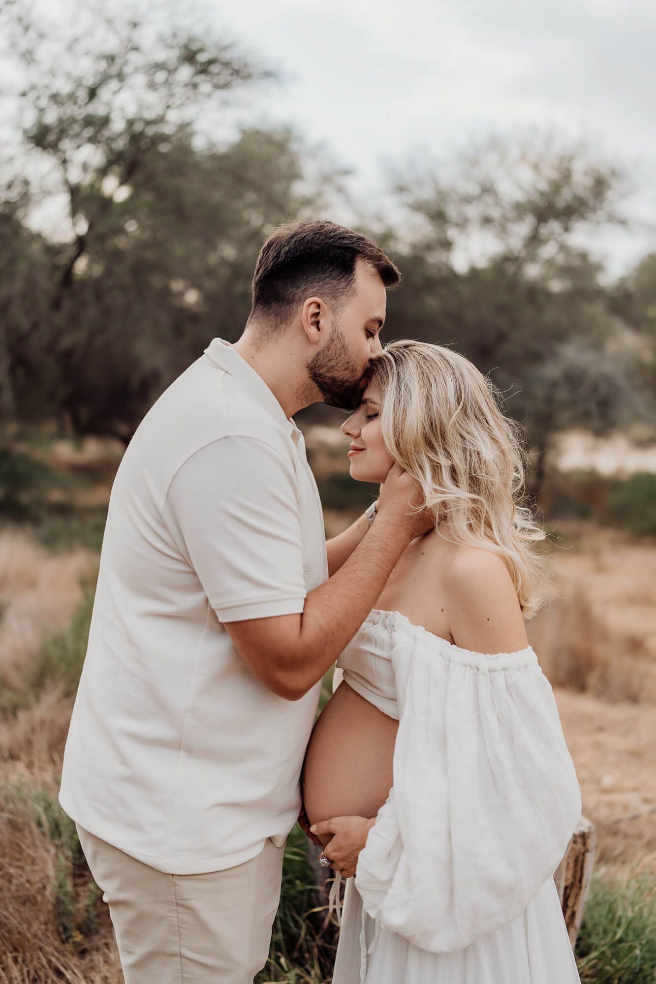 Intimate maternity moment captured in desert landscape with couple in cream colored outfits.