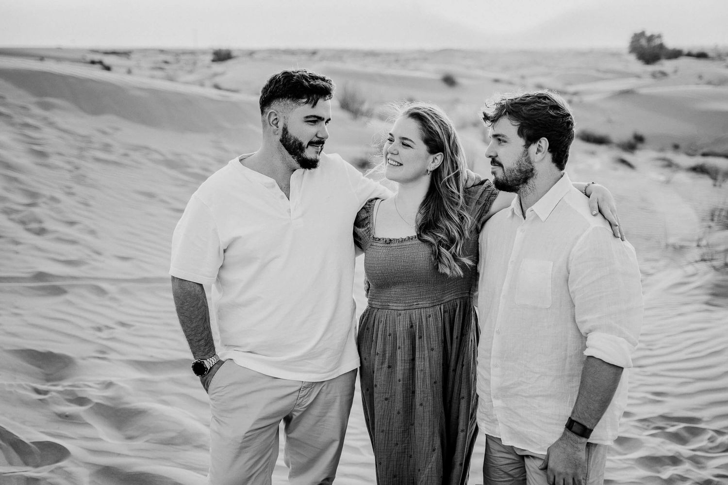 Black and white portrait of three companions smiling together among desert sand dunes at sunset.