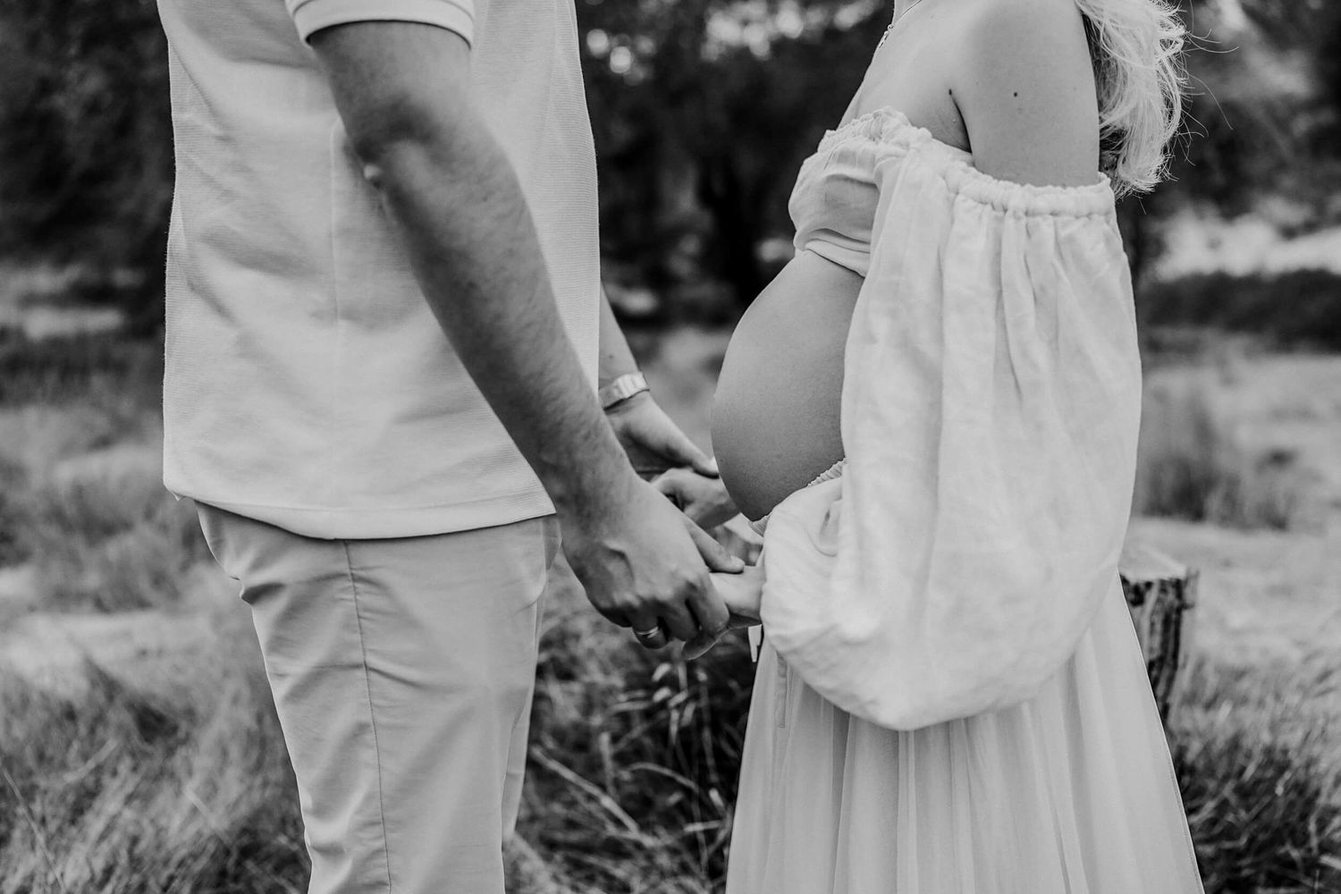 A couple holding hands in white clothing photographed in black and white against a natural outdoor backdrop.