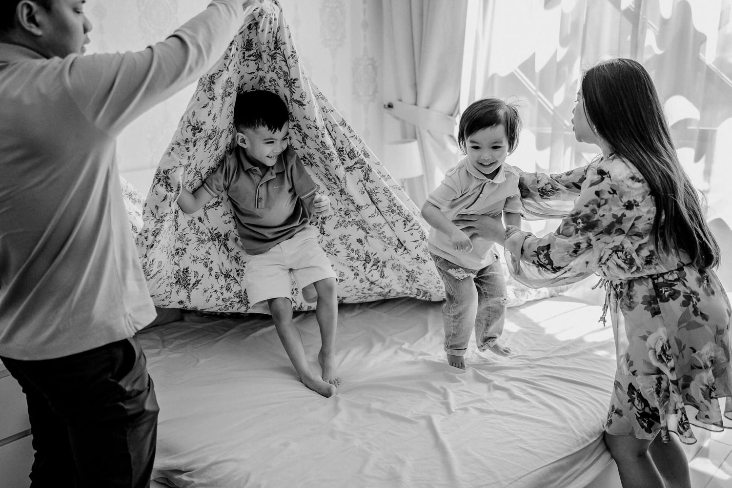 Black and white photo of people playing and having fun around a floral patterned tent in a bright room.