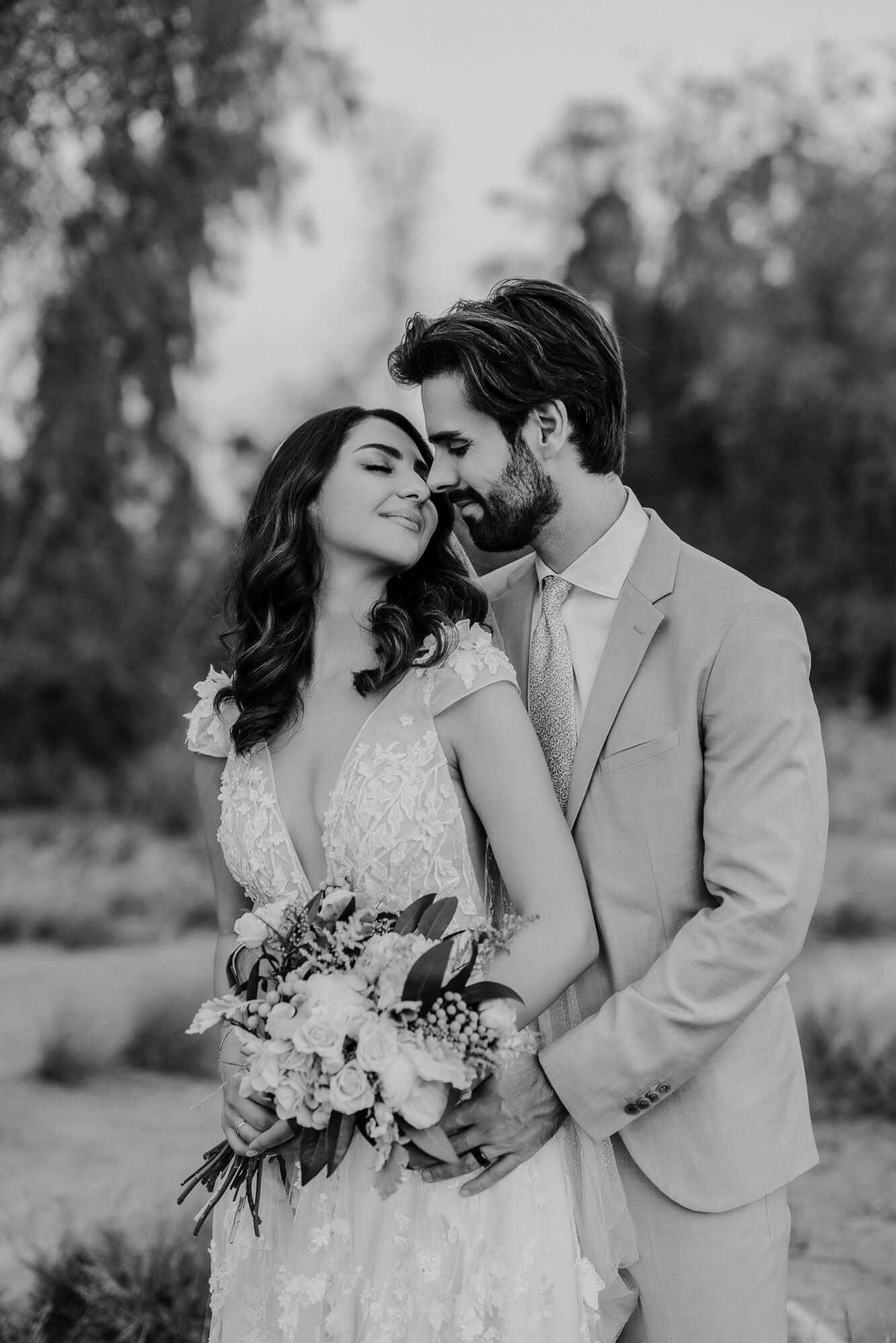 Black and white romantic wedding photo of couple embracing outdoors with bride holding floral bouquet.