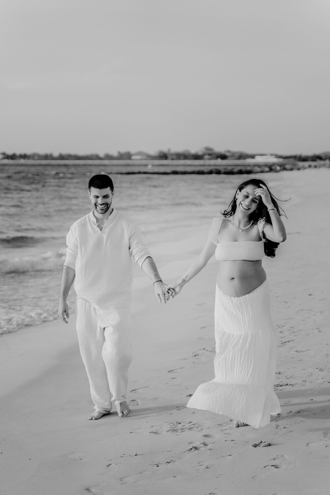 A couple in white outfits holding hands while walking on a beach during an overcast day.