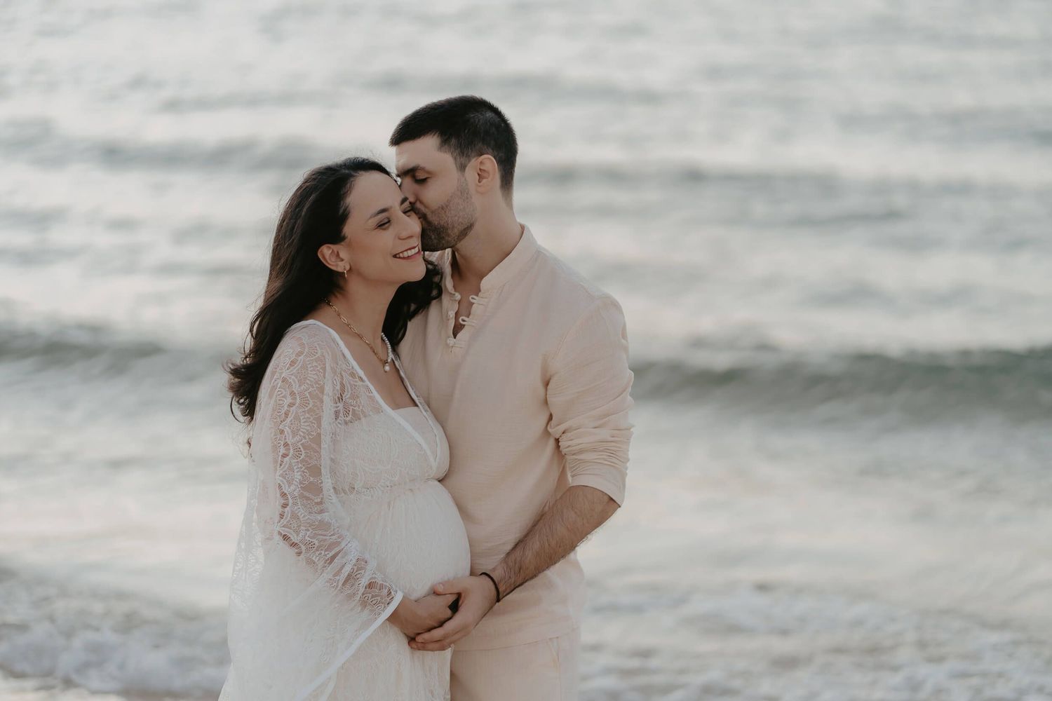 A romantic maternity photo session at the beach with a couple embracing against the soft ocean waves in the background.