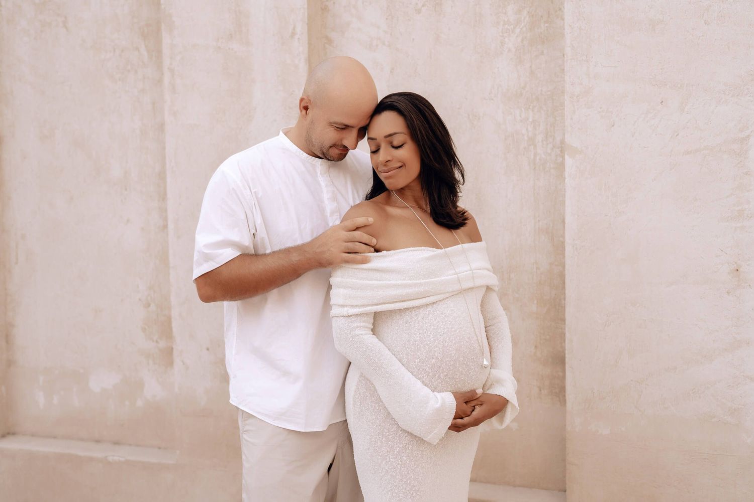 A tender moment between an expecting couple in white clothing against a neutral wall with architectural details.