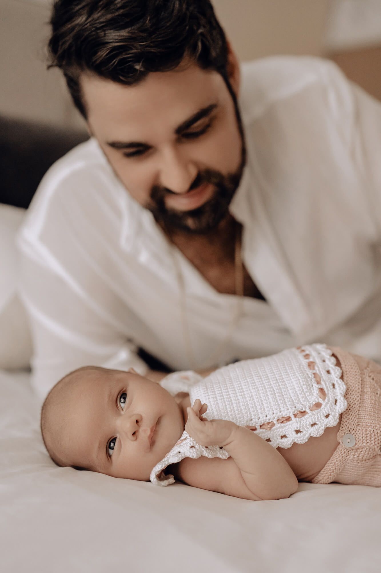 Parent gazes lovingly at newborn baby lying on white bedding.