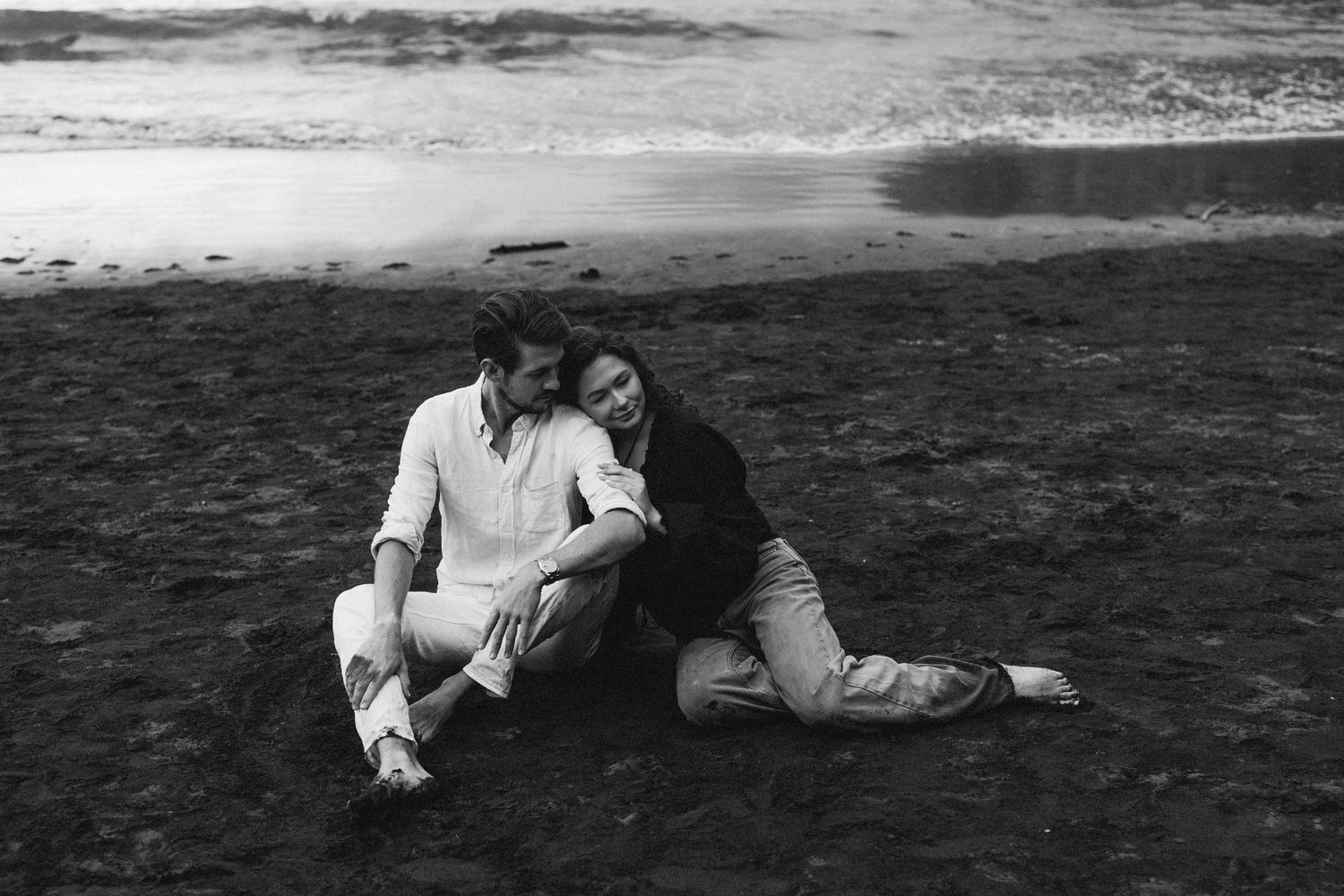 A couple sits intimately together on a dark sandy beach in a romantic black and white photograph.
