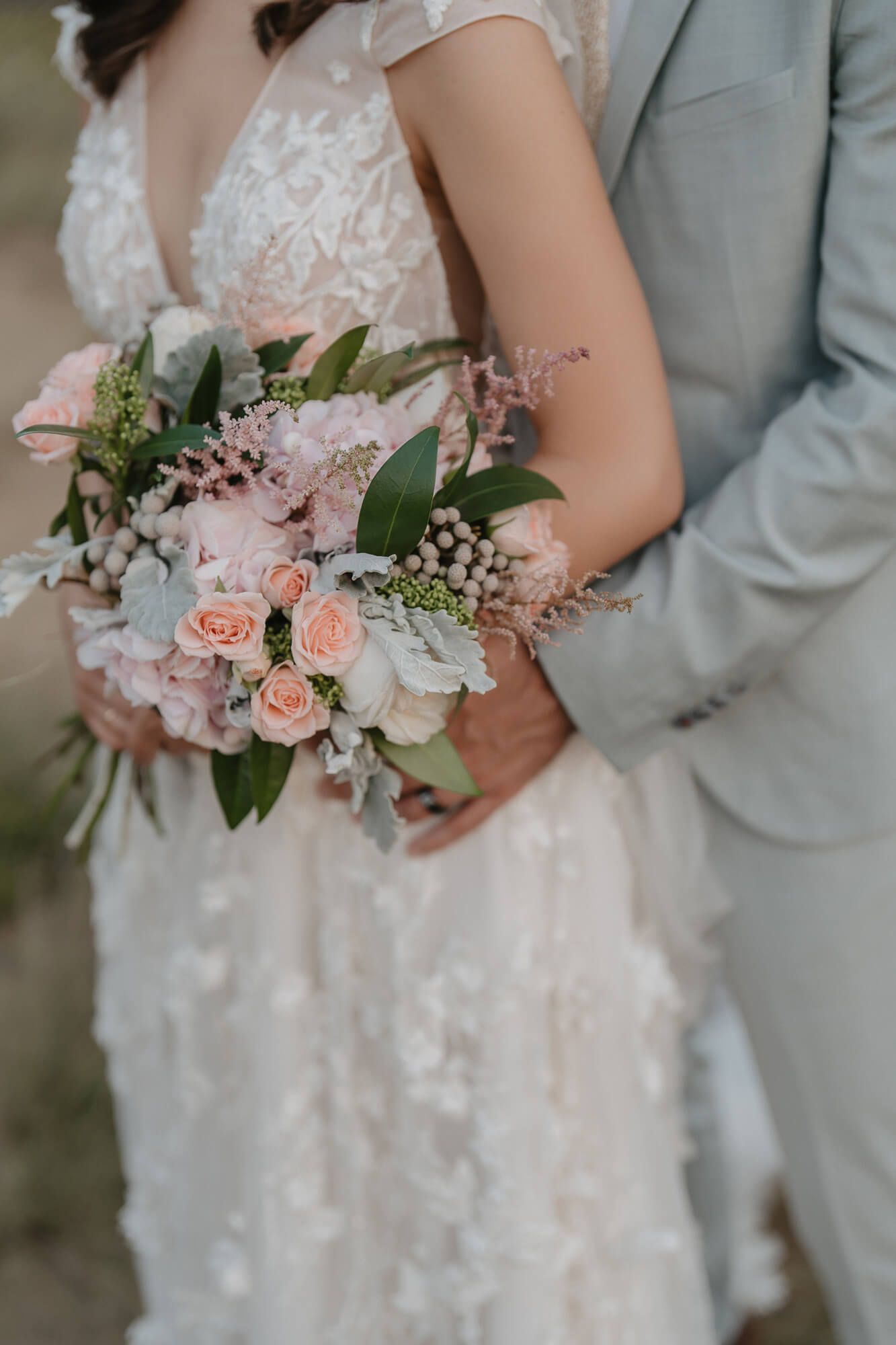 A romantic wedding bouquet of blush roses and mixed flowers held between a bride in lace and groom in grey.