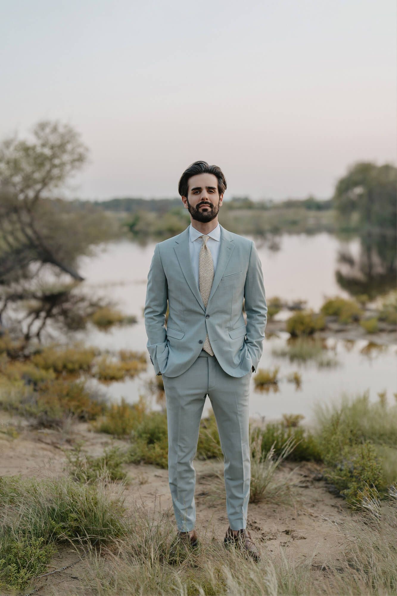 Person in light blue suit and hat poses in rustic outdoor setting near water during golden hour.