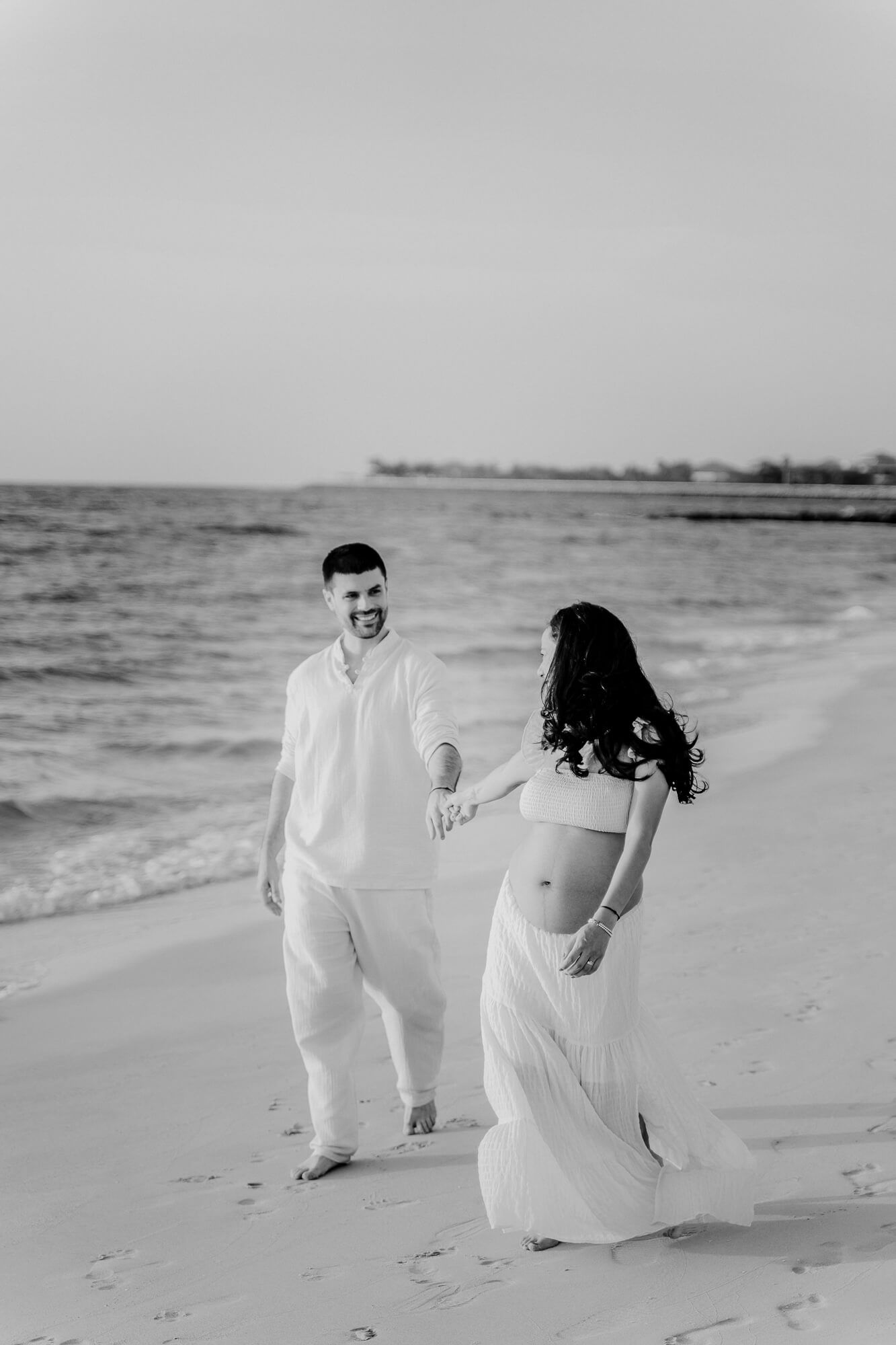 A couple in flowing white outfits walks together on a beach in black and white photography.