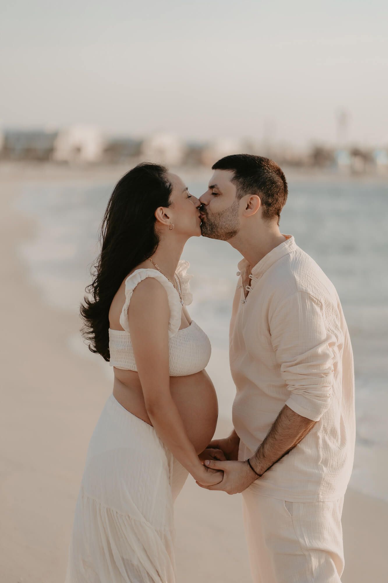 A romantic couple shares a tender moment together on a beach at sunset in white clothing.