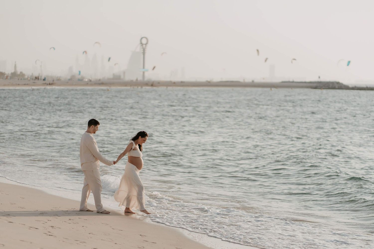 Two people in light colored attire walk hand in hand along a peaceful beach shoreline during golden hour.
