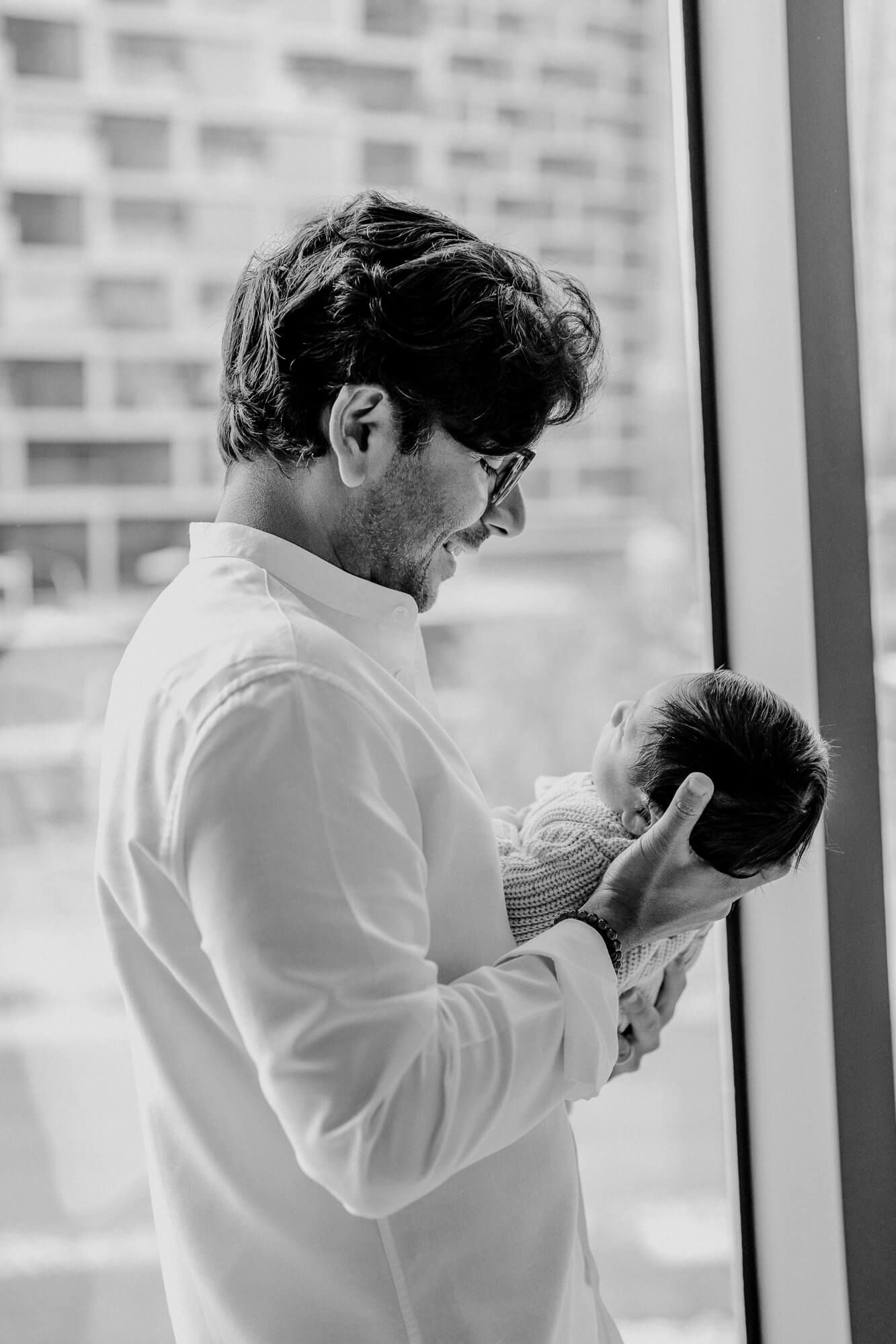Black and white portrait of parent cradling newborn by window.