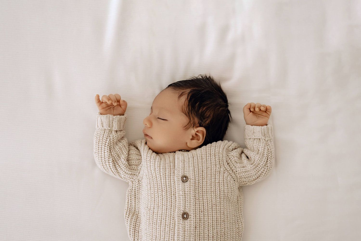 Sleeping newborn wearing cream knit sweater lying on white bedding in peaceful slumber.