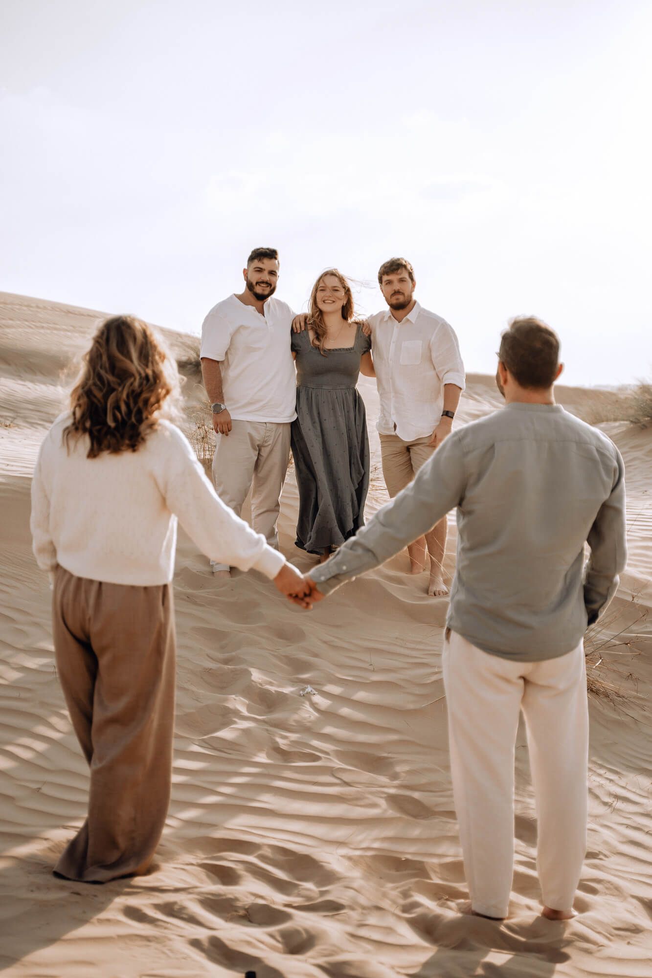 Wide shot of people holding hands in a circle on sandy dunes during sunset.