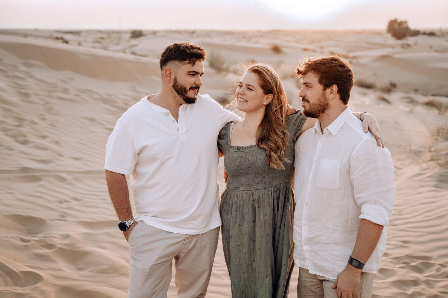 Three friends share a candid moment while posing together in desert sand dunes at golden hour.