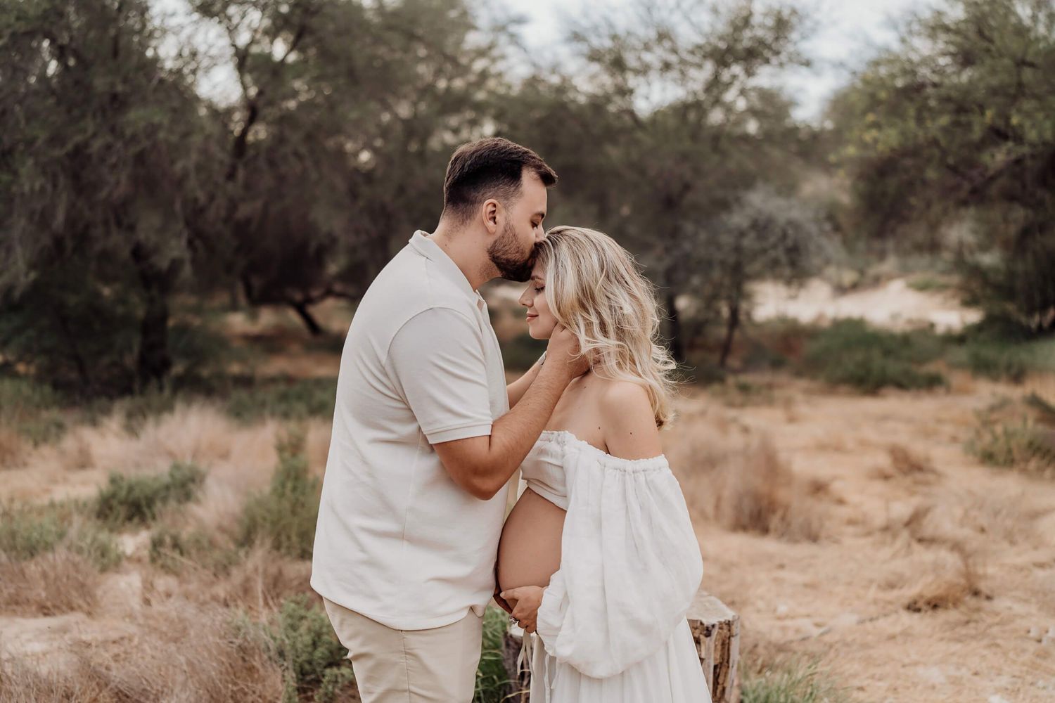 Desert maternity photography featuring couple in soft whites against natural landscape.