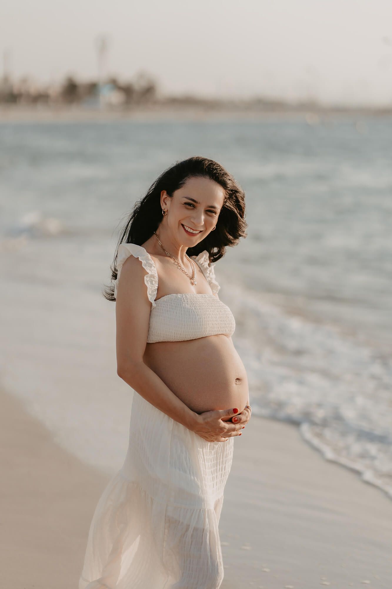 A person in a white lace dress poses on a beach at sunset while cradling their pregnant belly.