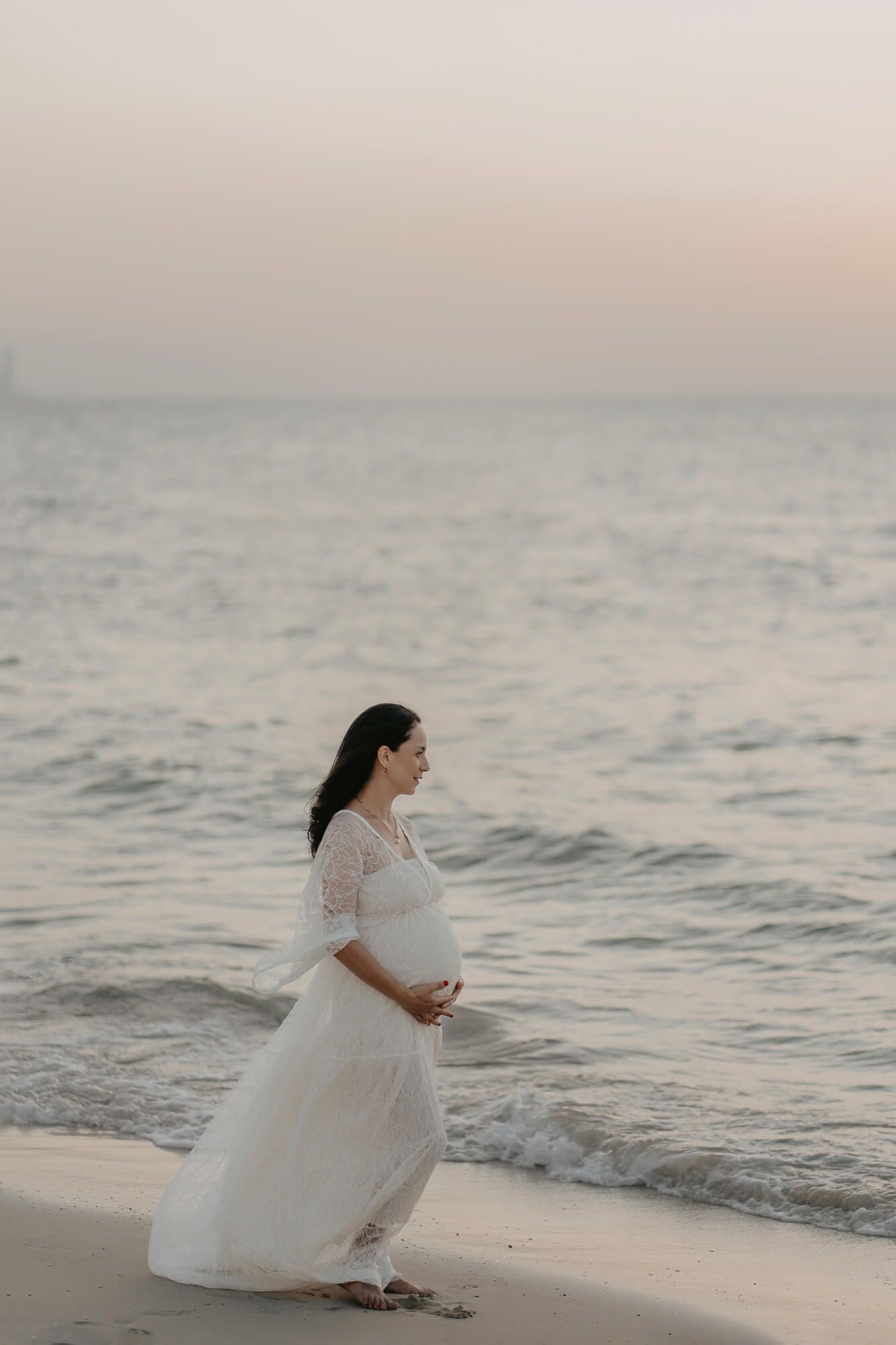 A pregnant woman wearing a flowy white dress stands on a serene beach at sunset capturing a peaceful coastal moment.