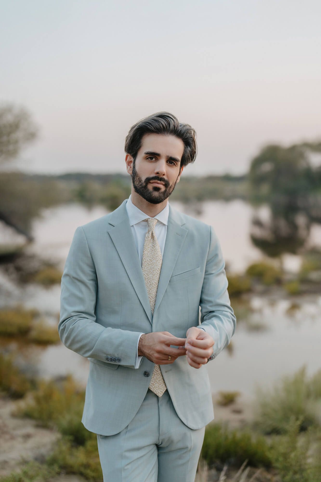 Someone wearing a light blue suit and yellow tie stands in front of a waterfront landscape at sunset.