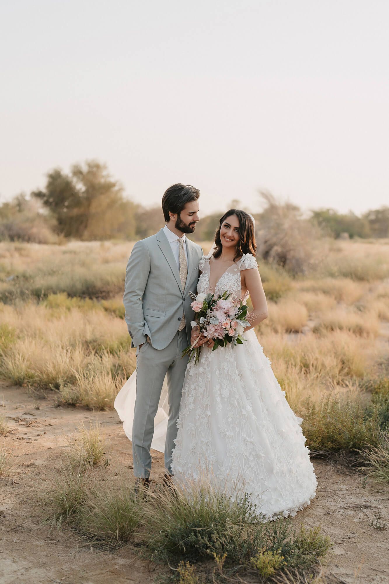 A couple in wedding attire poses together against a desert landscape with golden grasses at sunset.
