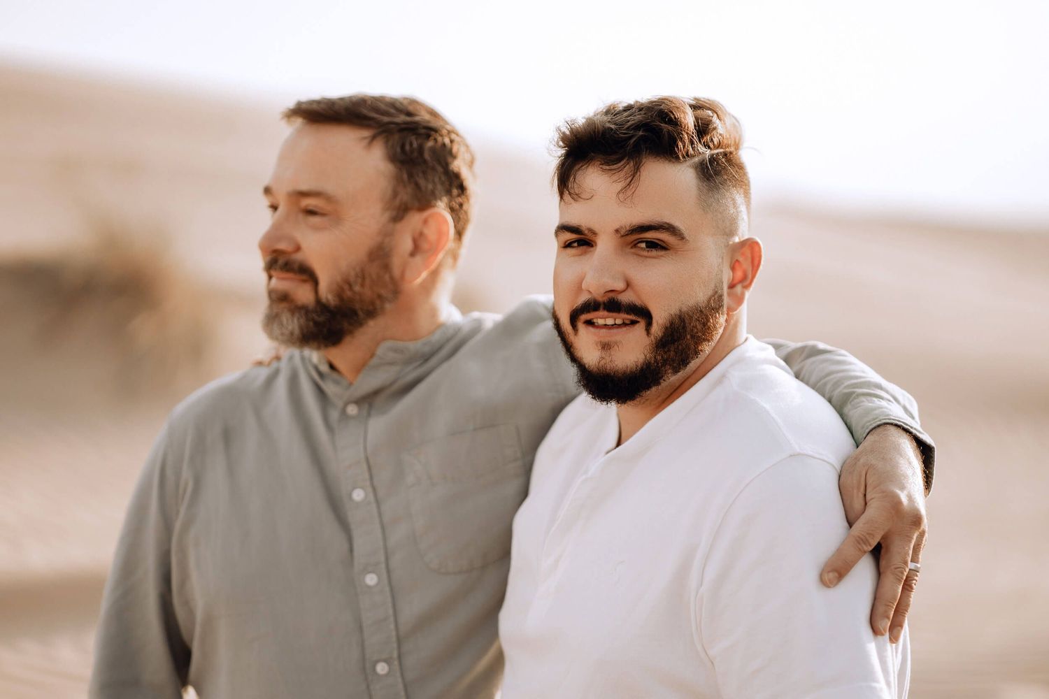 Two people in casual attire embrace while standing against desert sand dunes in warm lighting.