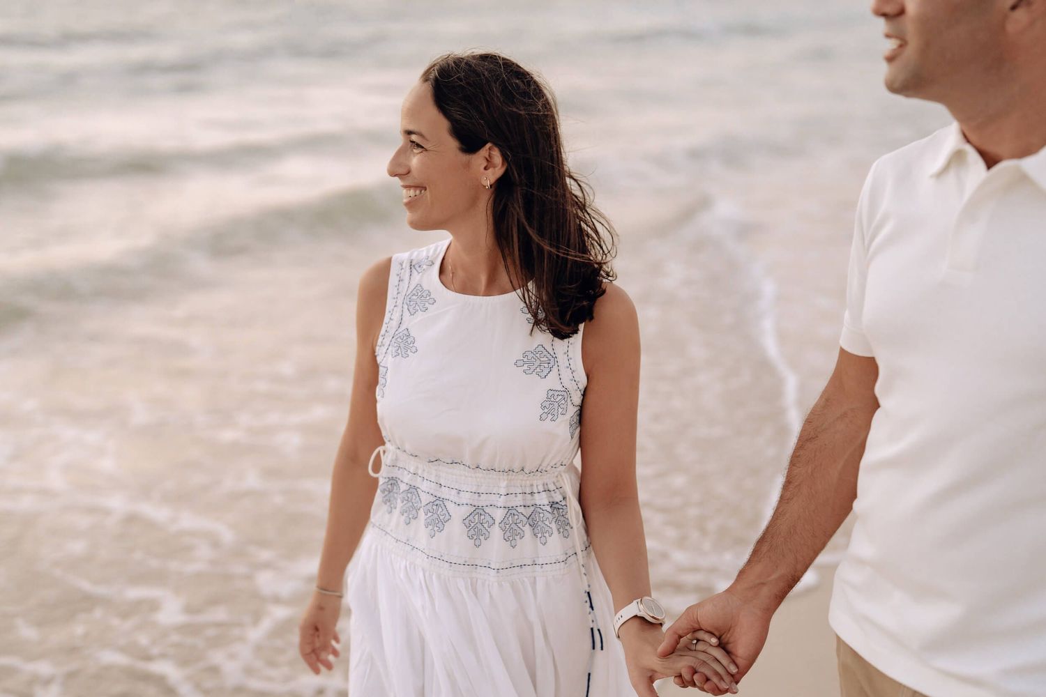 Side view of couple walking hand in hand on beach wearing white summer outfits.