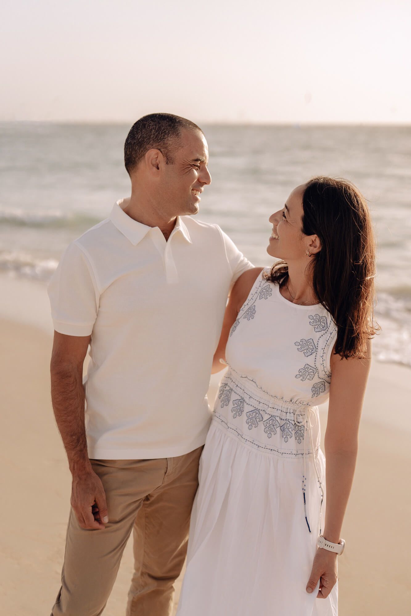 A couple in white and beige outfits share a loving moment on a beach at sunset.