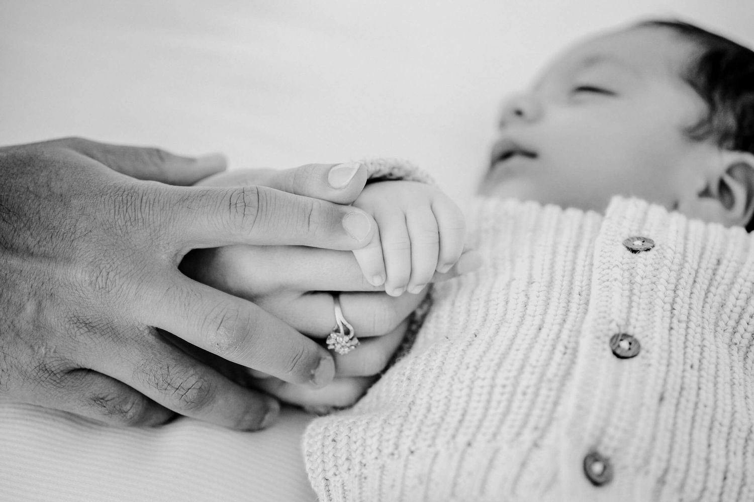 Black and white image of a sleeping infant holding a tender hand in a white knitted cardigan.