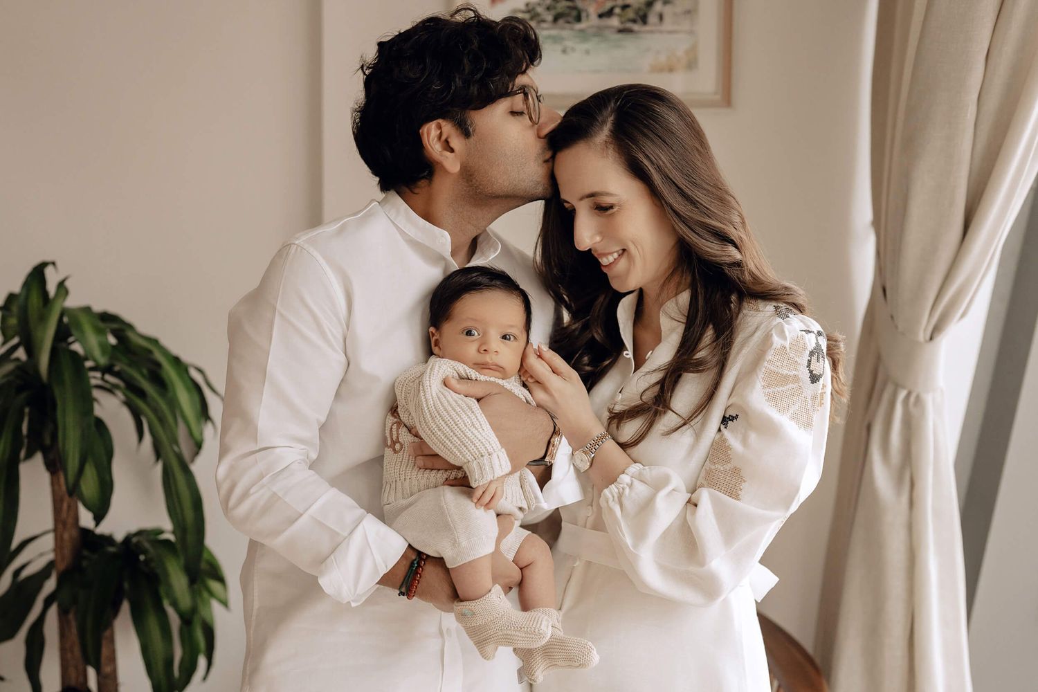 Parents in coordinated white outfits share loving moment with their infant beside window.