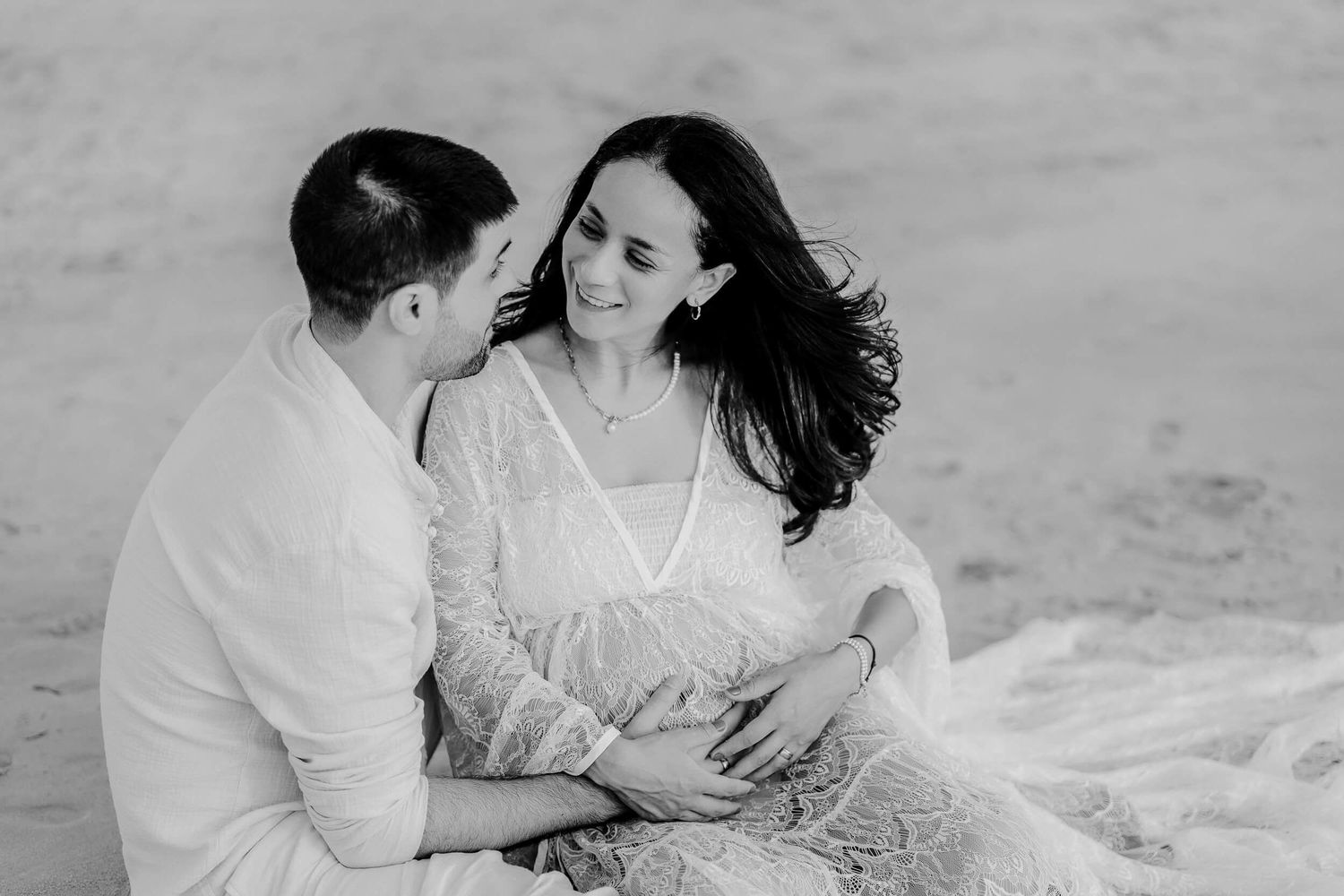 A couple in white clothes shares an intimate moment together on a beach during a black and white photoshoot.