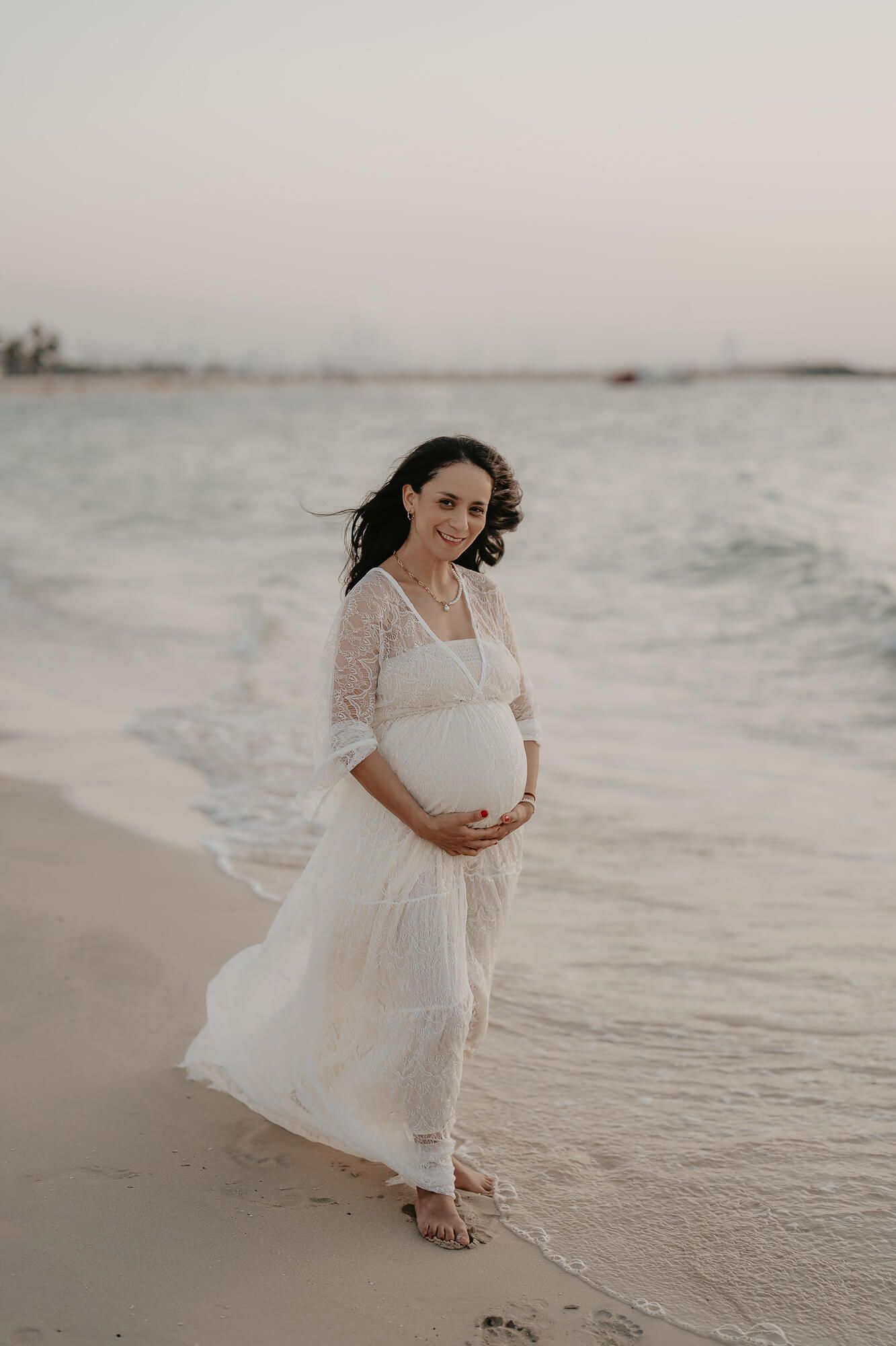 A beach photoshoot with an expectant mother in a flowing white lace dress walking along the shoreline at sunset.
