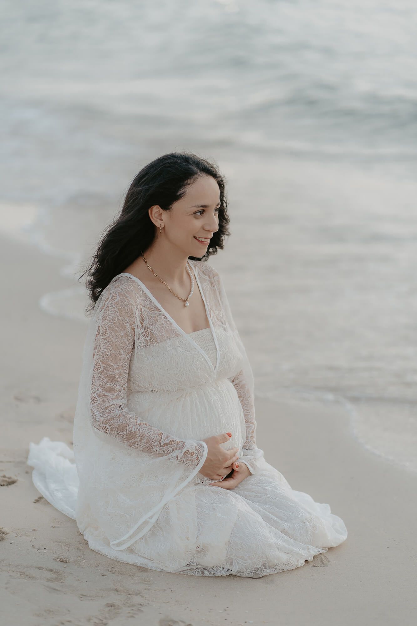 A serene maternity photo shows someone in a flowing white lace dress sitting peacefully on a beach at dusk.
