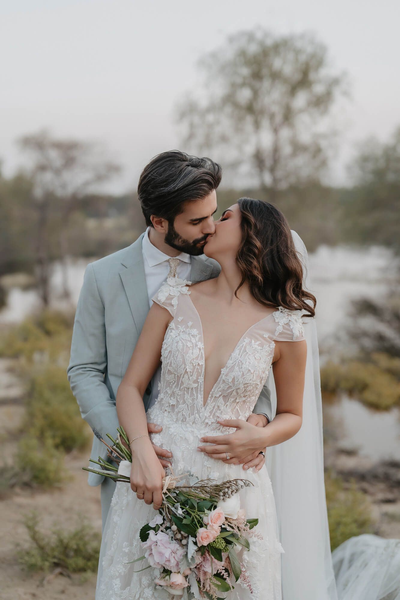 Romantic wedding couple sharing a kiss outdoors by a lake while holding a soft pink floral bouquet.