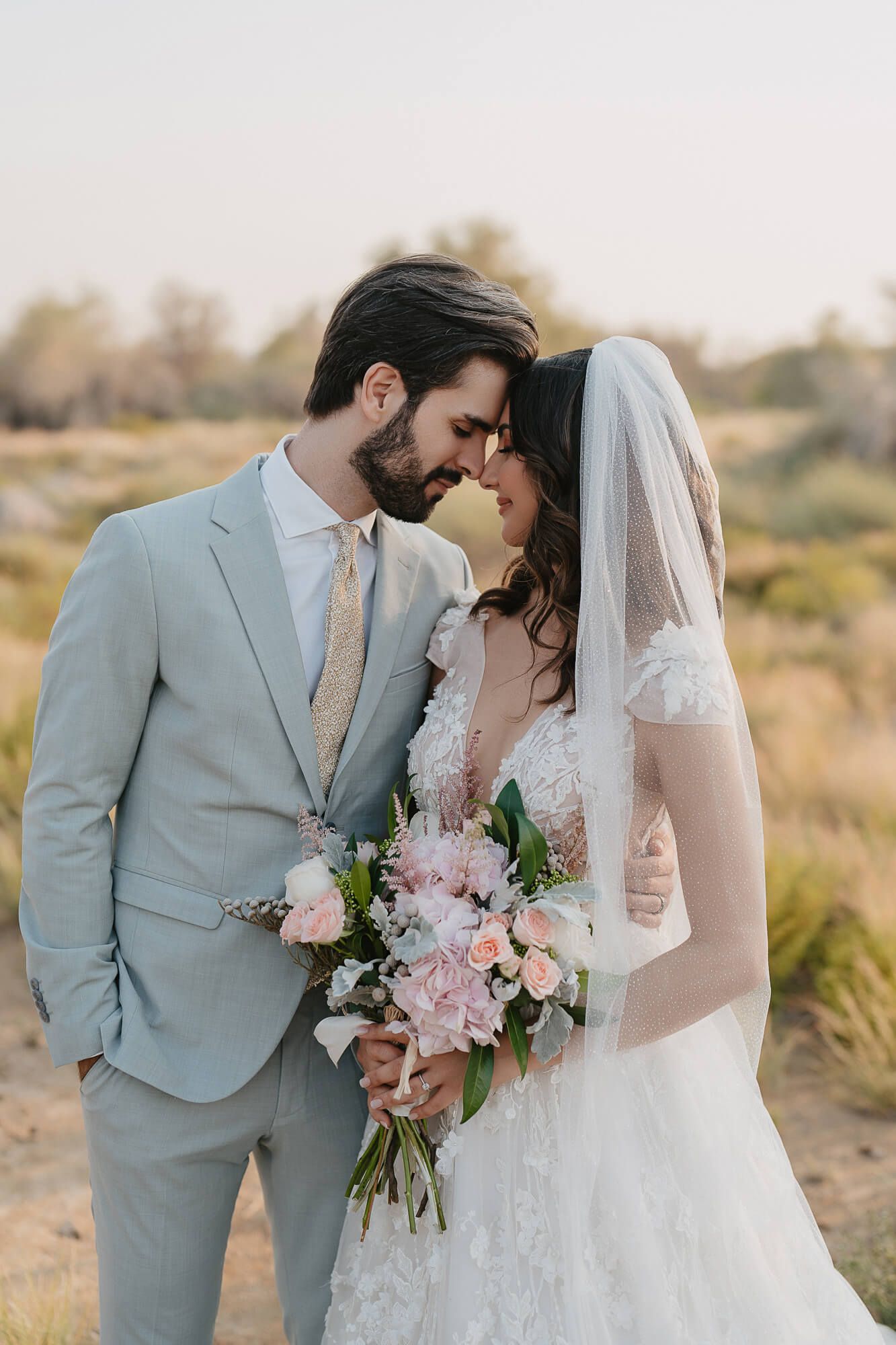 Bride and groom share an intimate moment at sunset in a desert setting with soft pink florals.