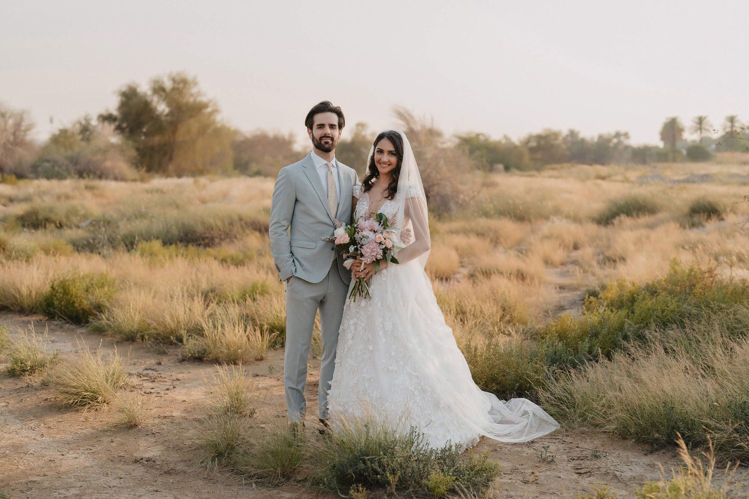 A newlywed couple stands together in a desert setting with dried grass and natural scenery.