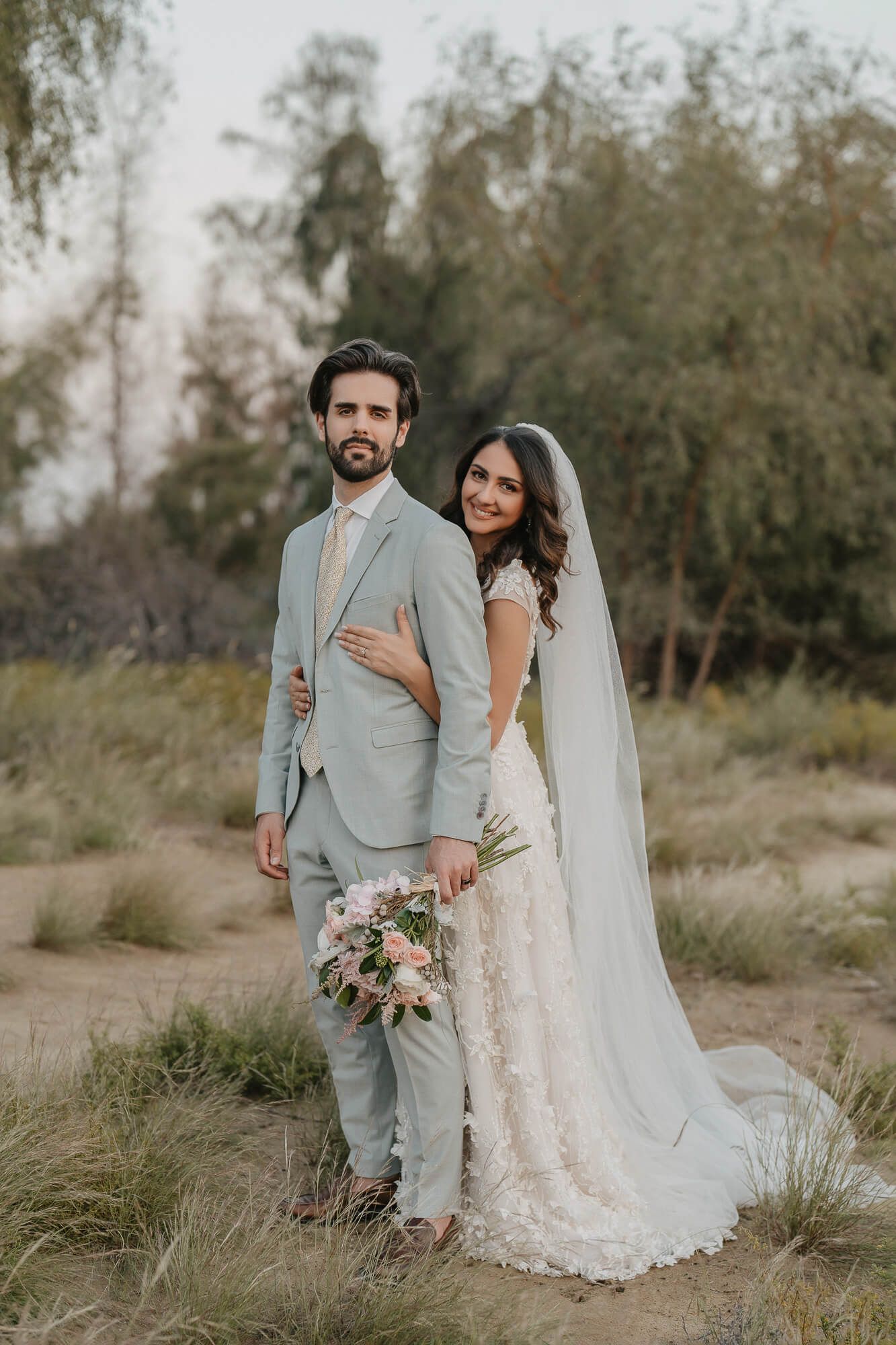 Newlyweds pose together outdoors with bride in lace gown and groom in light gray suit amid desert brush landscape.