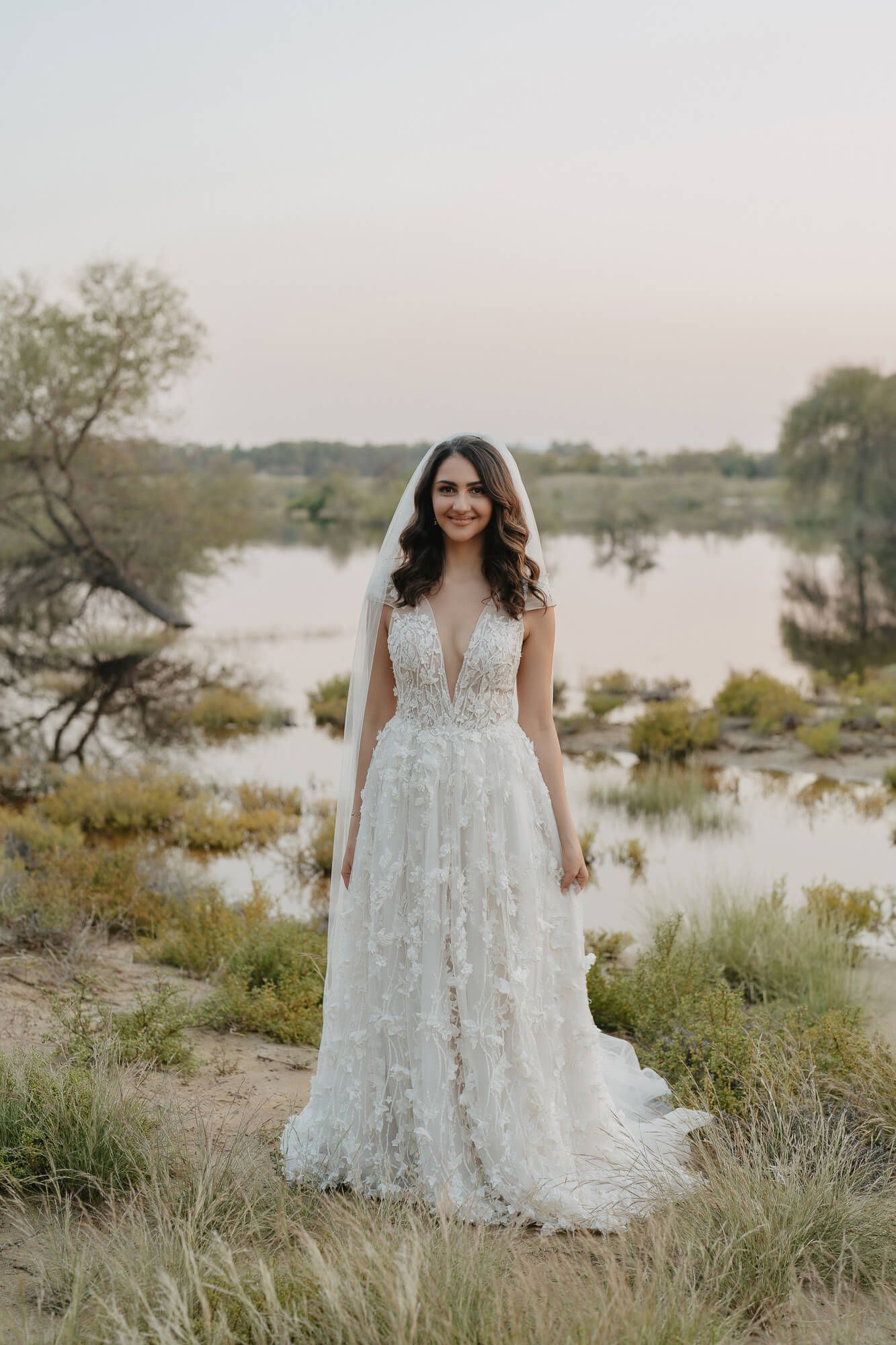 Ethereal white lace wedding dress with floral details photographed in a dreamy lakeside setting at sunset.